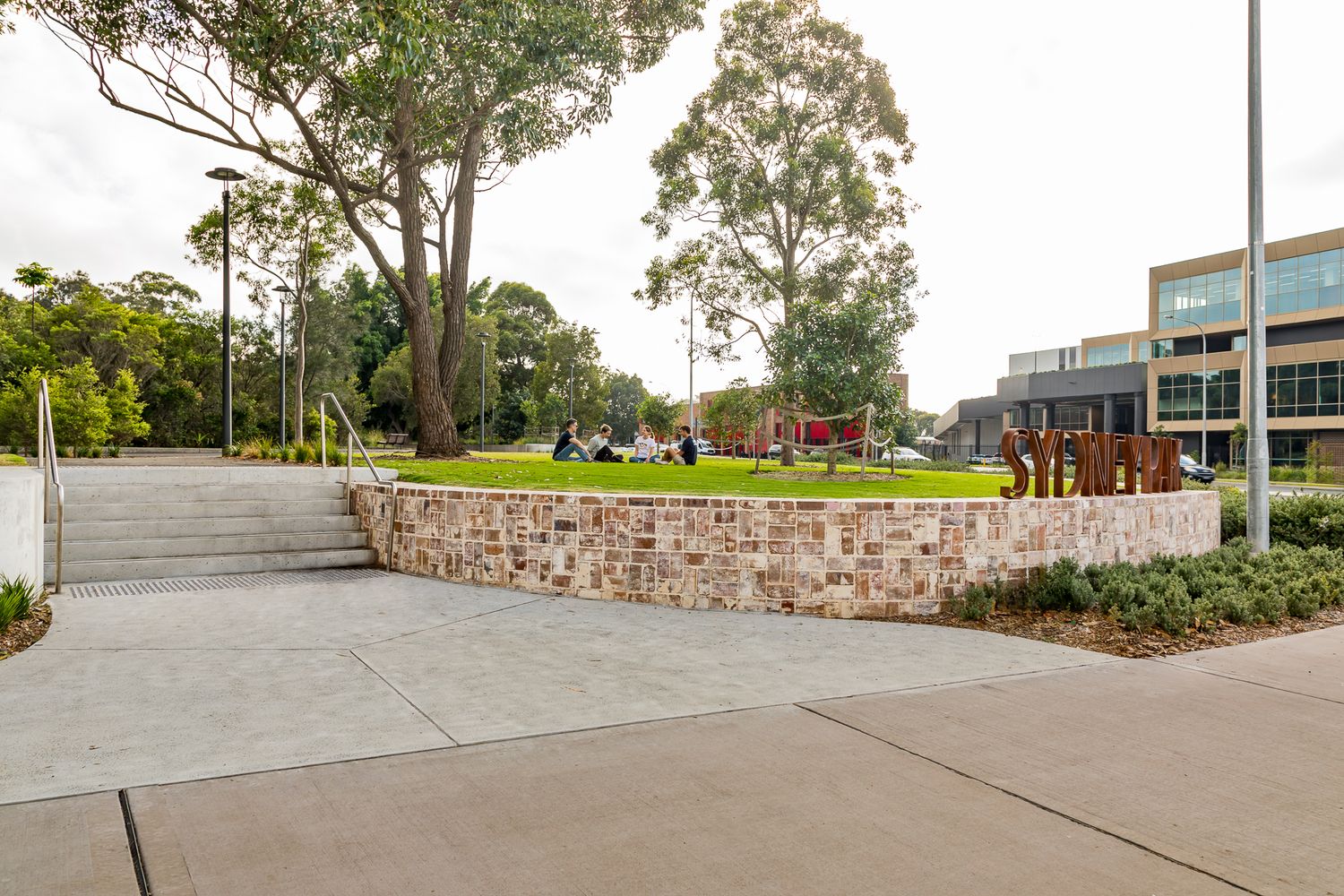 Sandstone retaining wall and concrete steps frame a green lawn with gum trees and people relaxing at Sydney Park entrance.