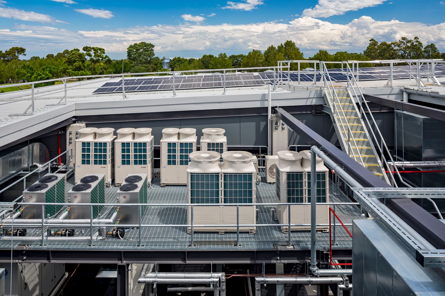 Rooftop plant area with air conditioning units, solar panels and steel stairs set against a blue sky with clouds.