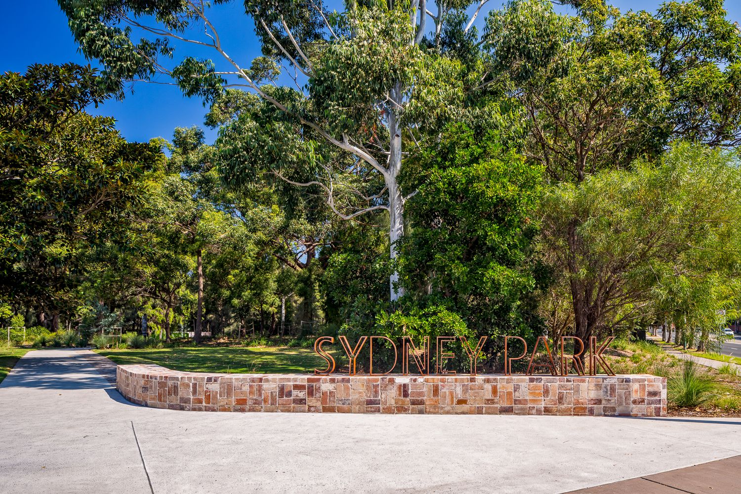 Sydney Park entrance sign on a curved stone wall beside a paved path, shaded by tall eucalyptus trees and bright blue sky.