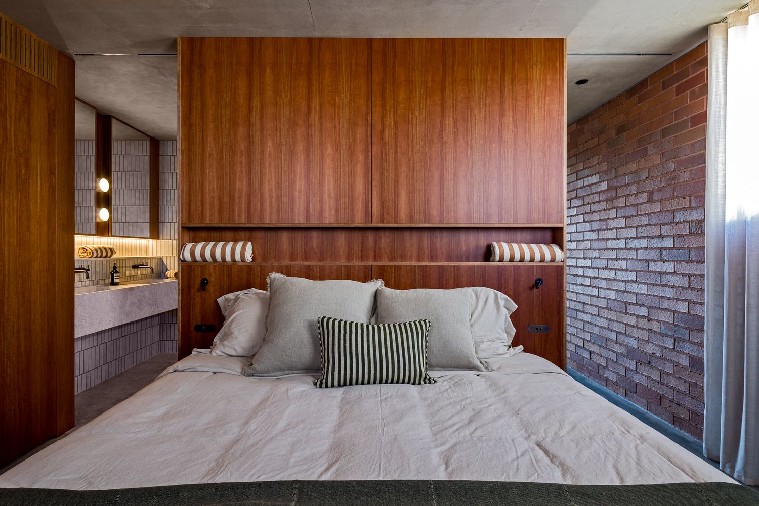 Bedroom with a timber headboard wall, exposed red brick, and a bed dressed in gray pillows and a striped cushion, with a bathroom beyond.