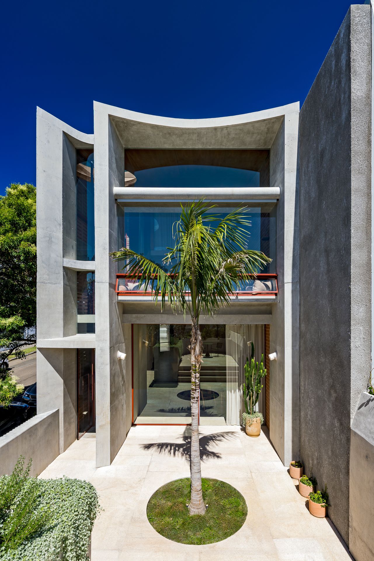 Symmetrical concrete house facade with curved parapets, a central palm tree, lawn circle and potted plants.