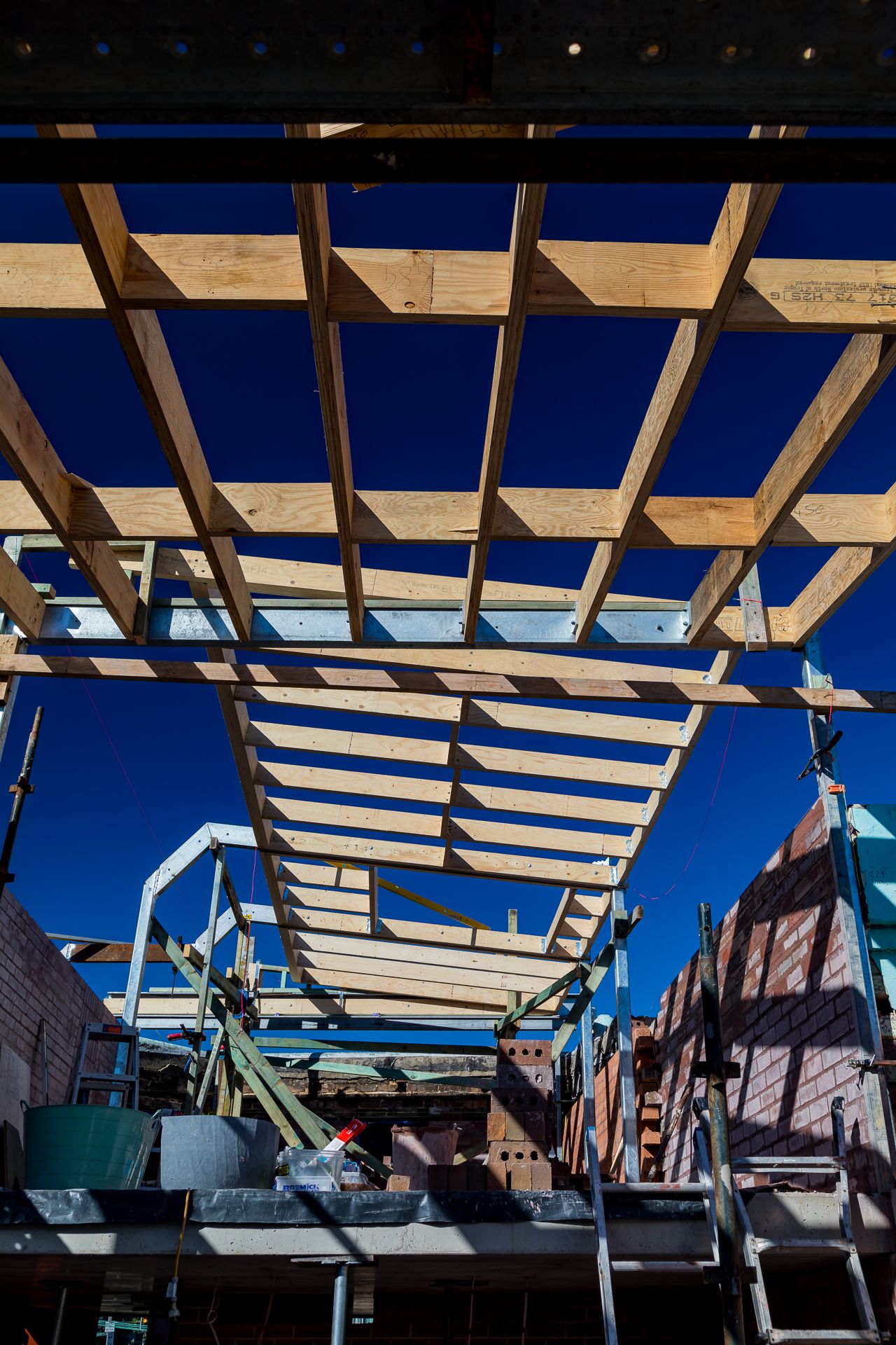 Timber roof rafters and steel supports seen from below under a blue sky at a brick house renovation site.