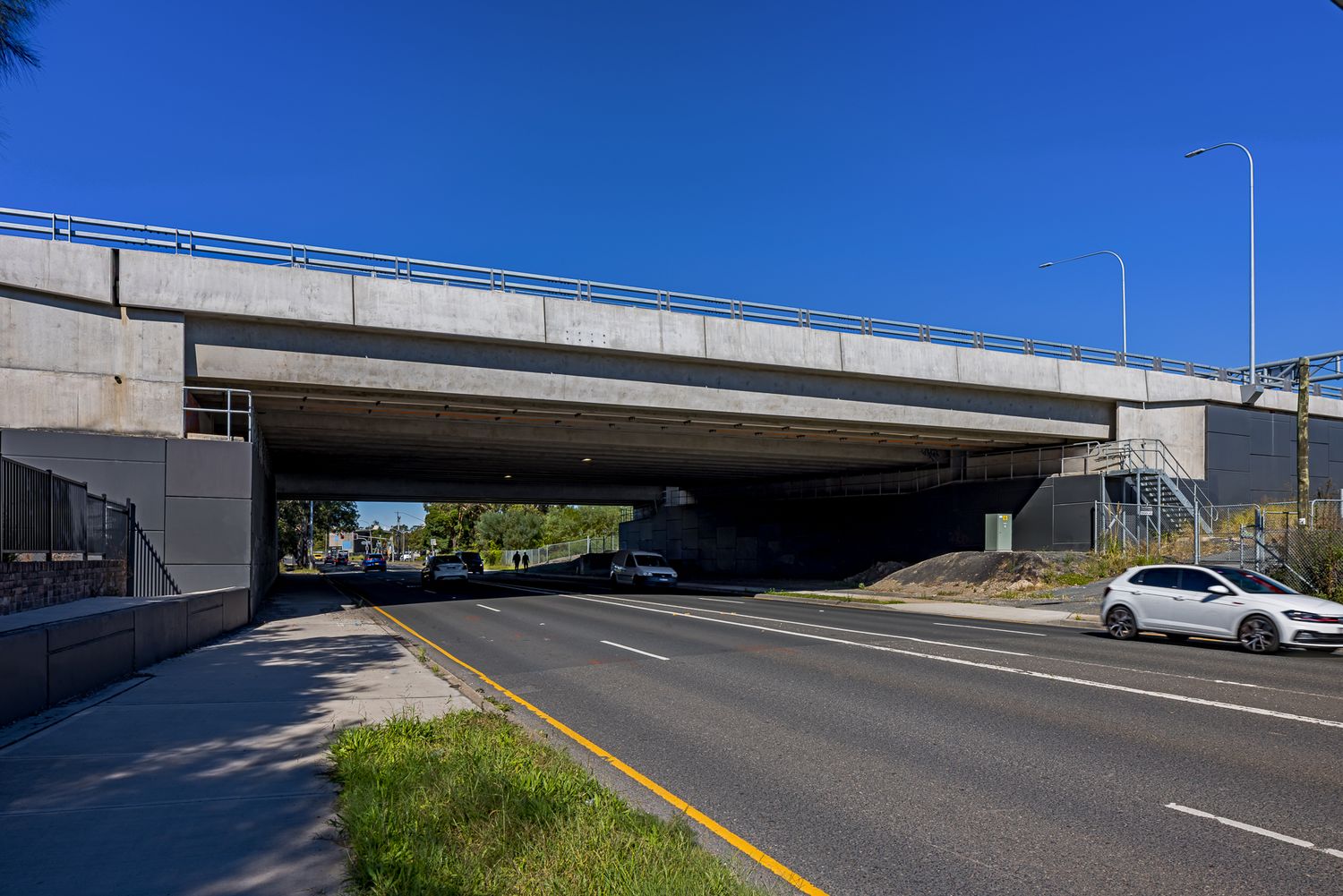 Concrete road overpass spanning a multi-lane road under a clear blue sky, with a white car driving beside a footpath and grass verge.