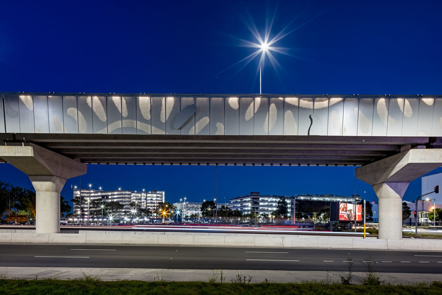 Elevated rail guideway with decorative patterned panels spans a city street at dusk, with illuminated buildings and a bright starburst light