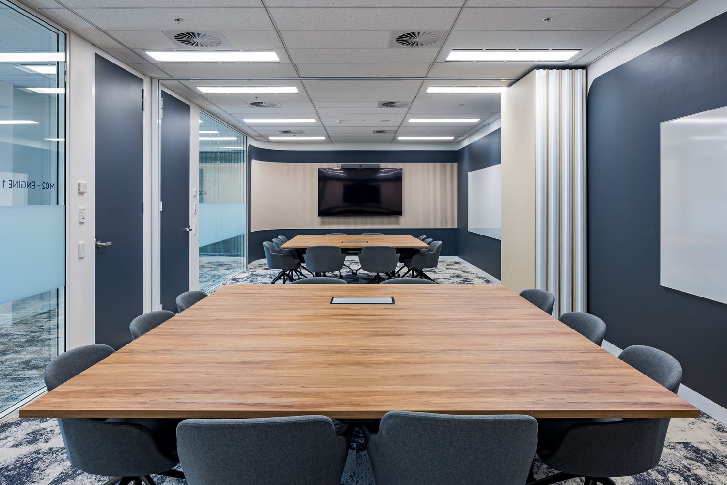 Spacious conference room with a long wooden table, gray chairs, a wall-mounted monitor and navy accent walls viewed through glass.