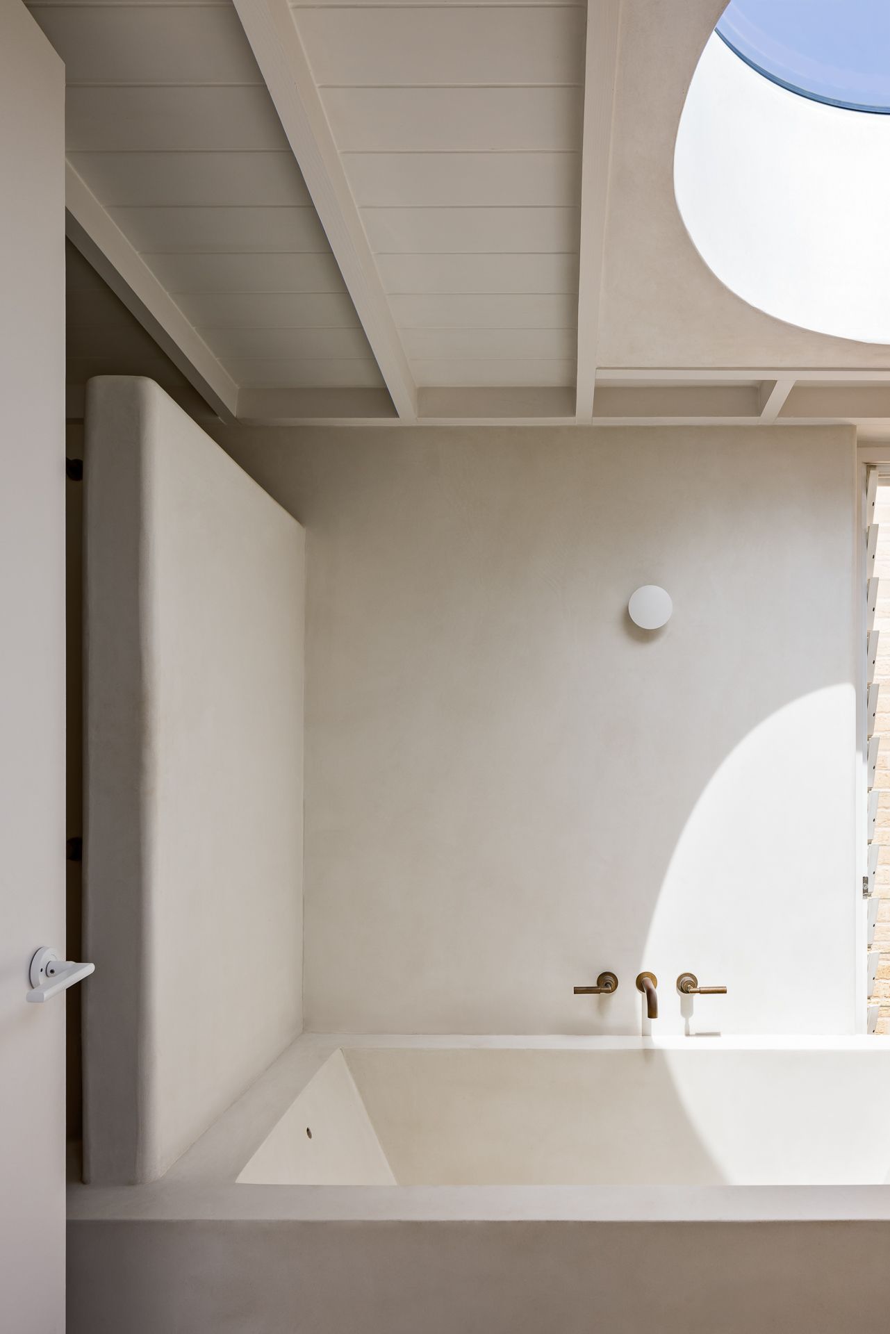 Sunlit bathroom corner with sculptural plaster tub, brass wall-mounted taps, white beams and skylight casting soft shadows.