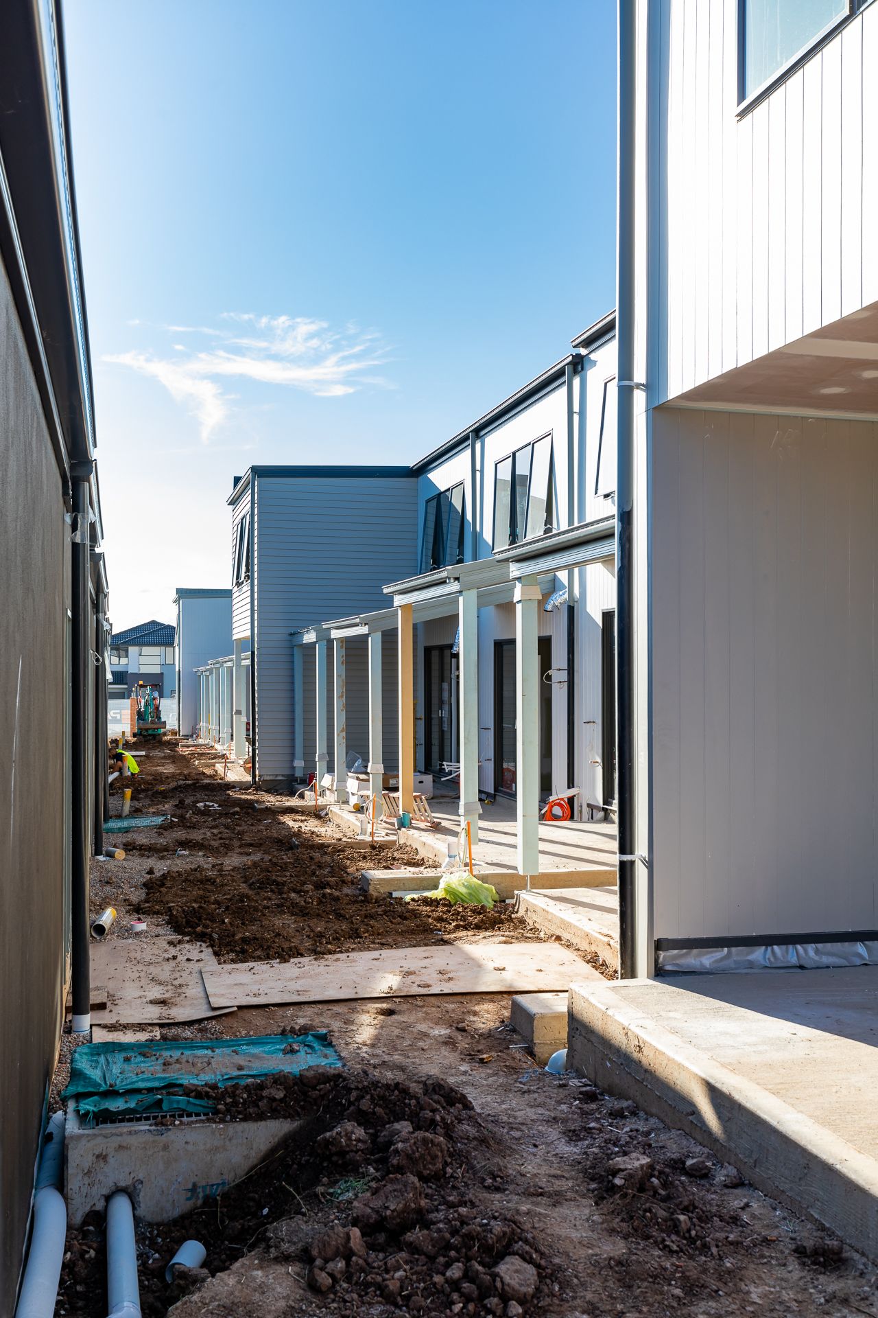 Narrow construction corridor between new modern townhouses with fresh soil, drainage pipes and covered porches under a blue sky.