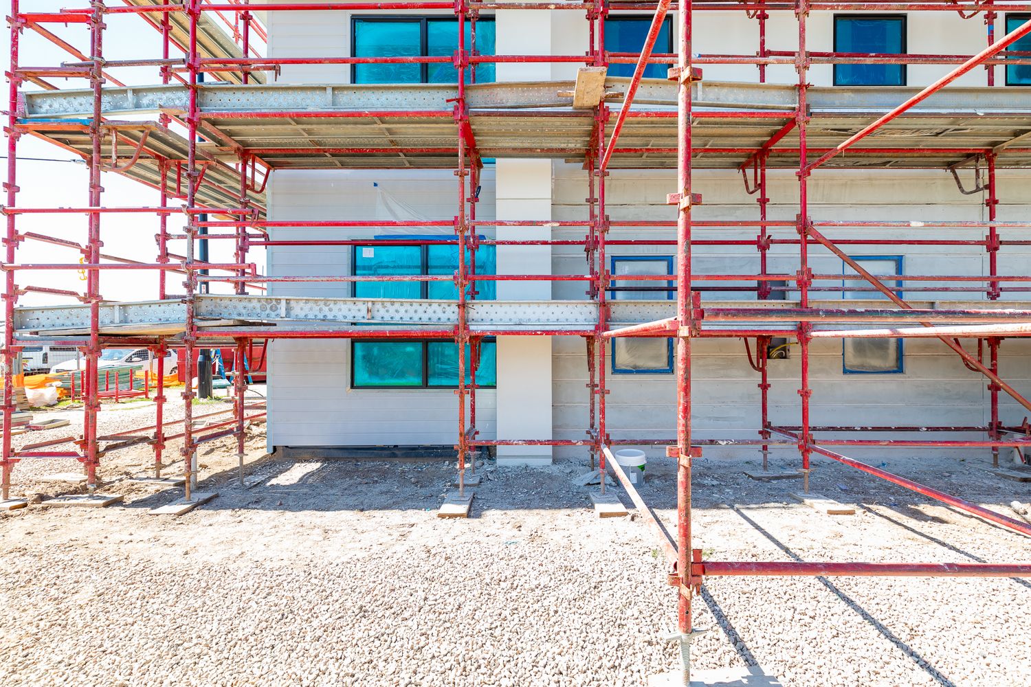 Red scaffolding covers the front of a white house under construction with blue-framed windows and a gravel ground surface.