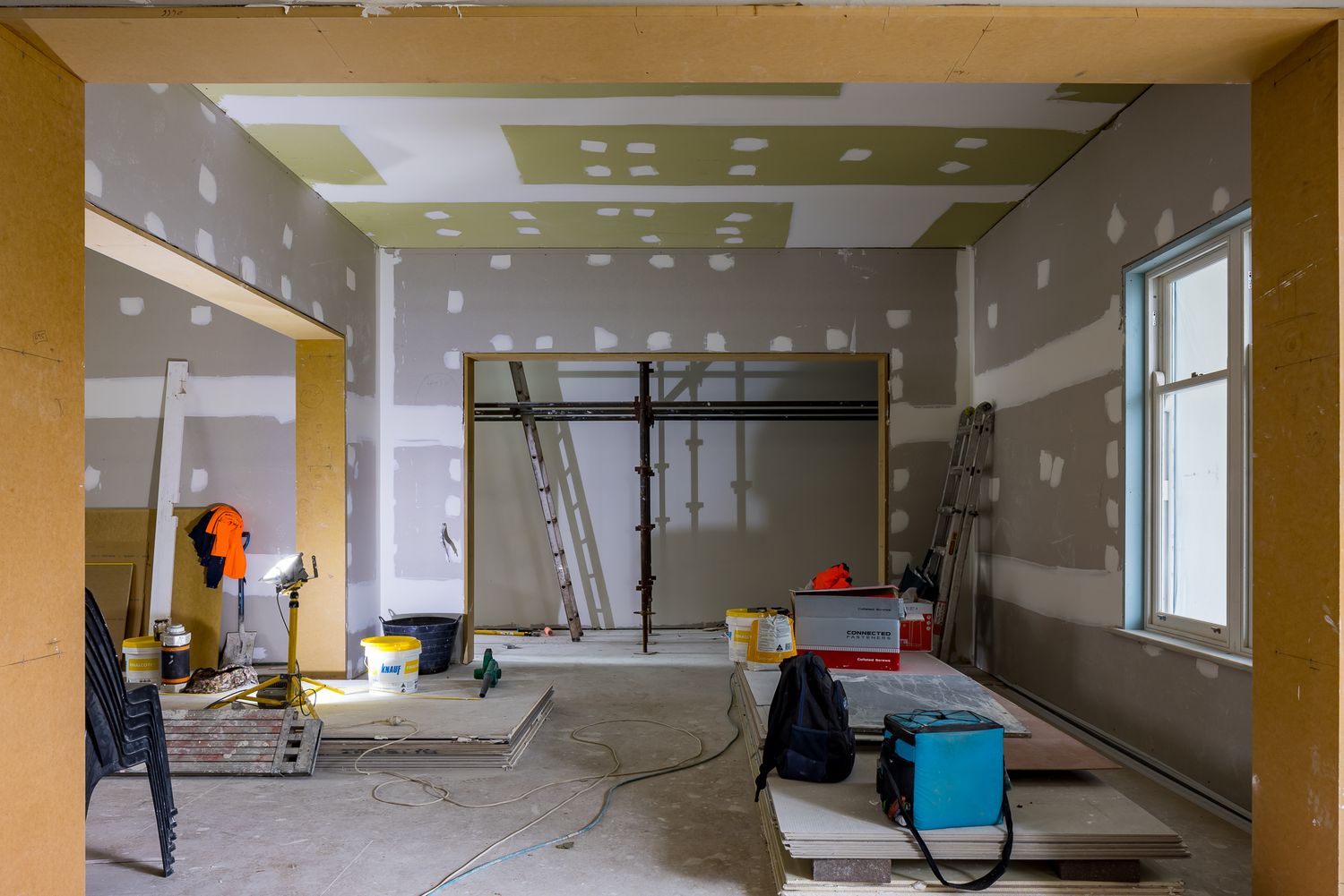 Home renovation in progress with exposed plasterboard walls, ladders, tools and building supplies scattered across a concrete floor.