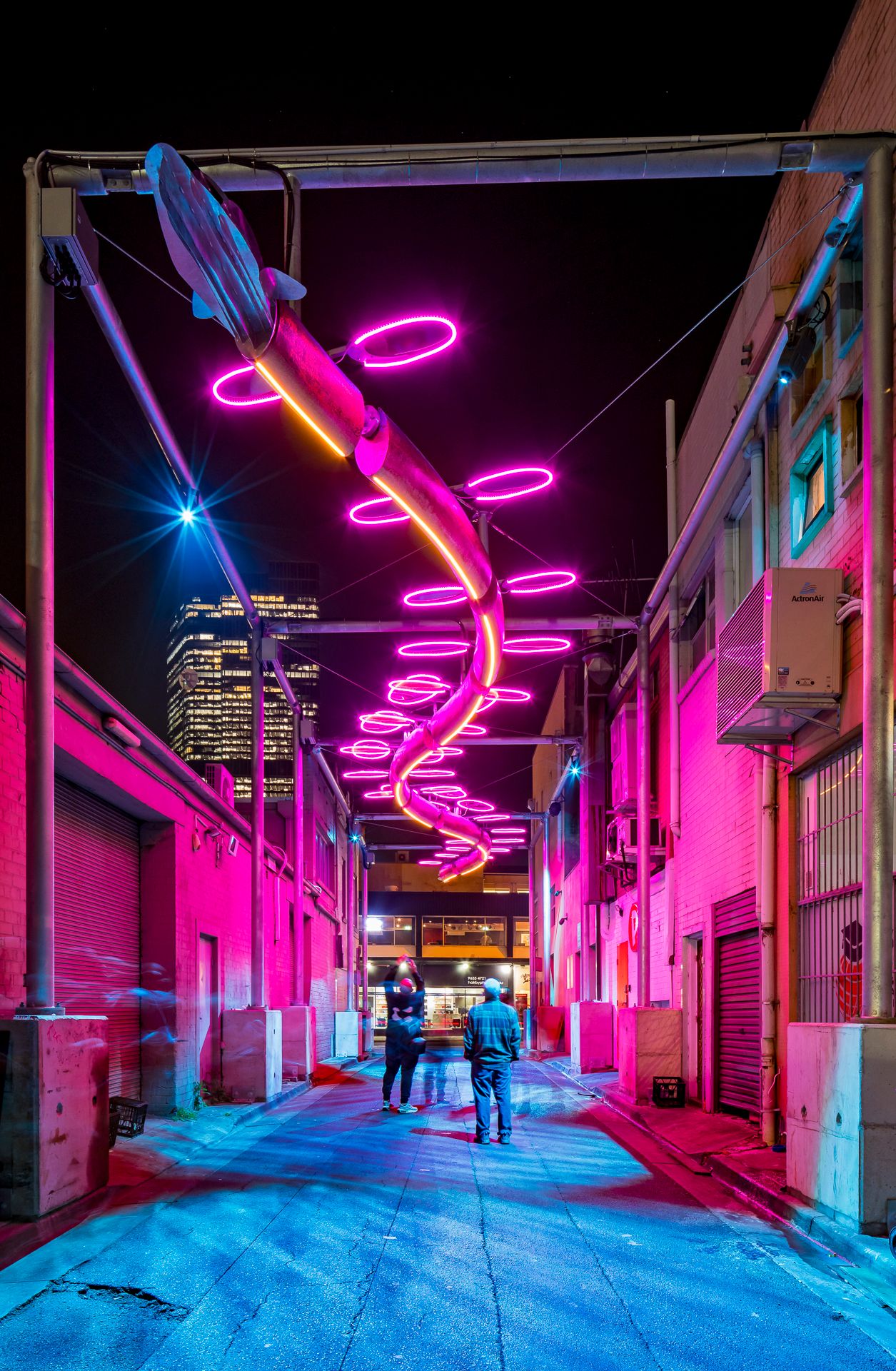 People walk through a nighttime laneway lit in vivid pink and magenta by a spiral neon art installation hanging overhead.