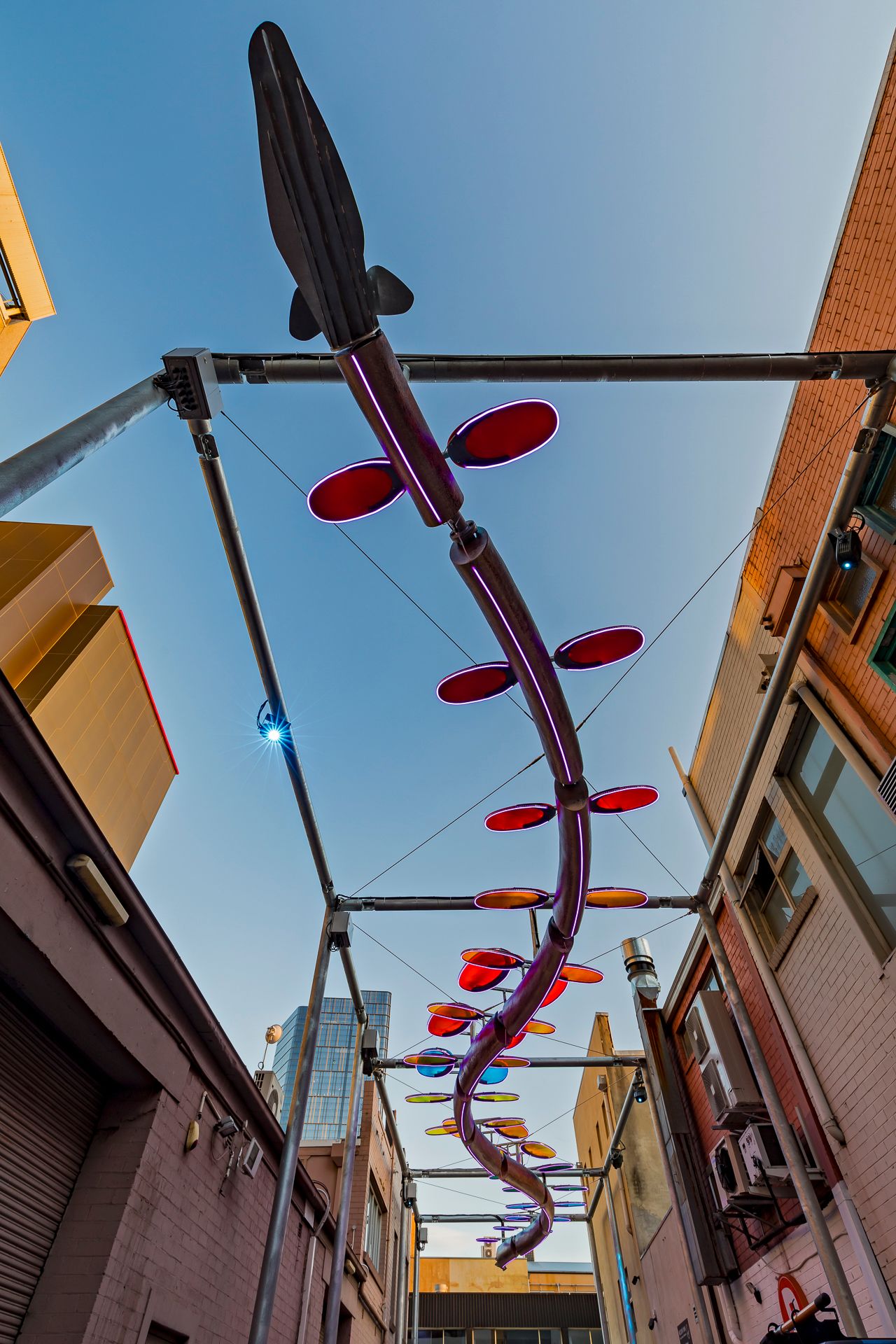Upward view of a fish-topped serpent sculpture with red oval discs suspended on a steel frame between city buildings.