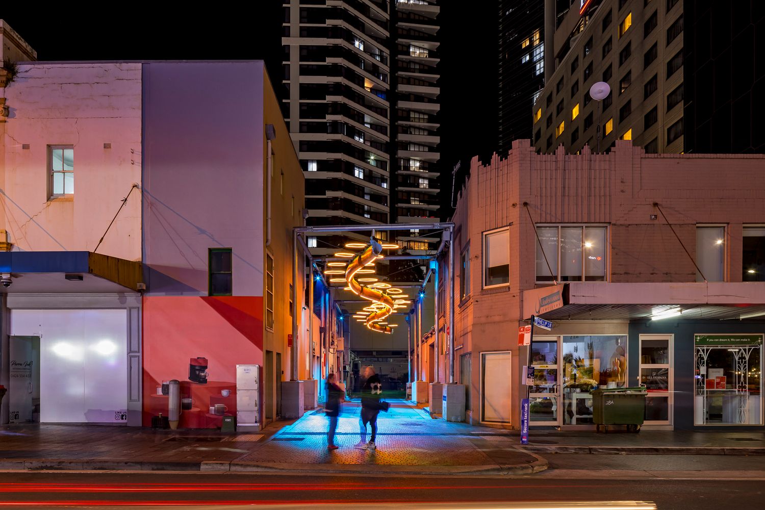 Golden orange neon spiral artwork illuminates a city alley at night as two pedestrians stand near blue-lit pavement.