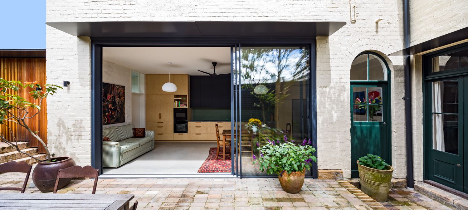 Renovated brick home with open sliding glass doors connecting a bright living area to a sunny paved courtyard with potted plants.
