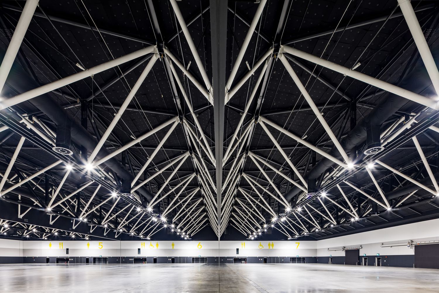 Symmetrical view of a large exhibition space ceiling with dramatic angular white steel trusses radiating over a polished floor.