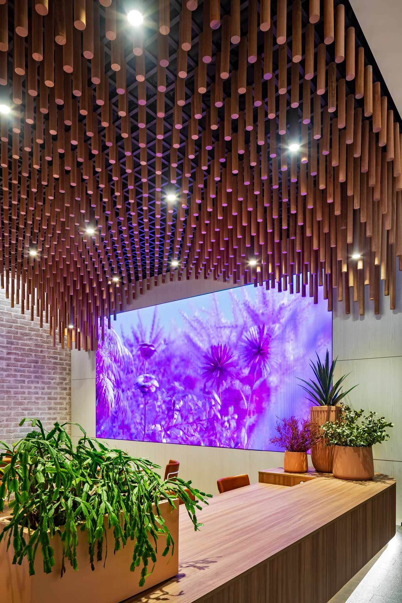 Modern lounge with hanging timber dowel ceiling, large purple floral video screen, wooden table, and potted plants.