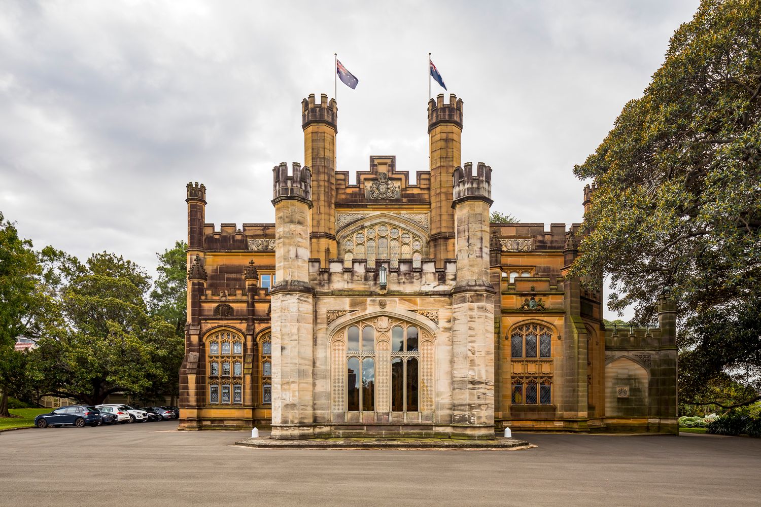 Gothic Revival sandstone building with battlemented towers and flags, with a parking area and mature trees in front.