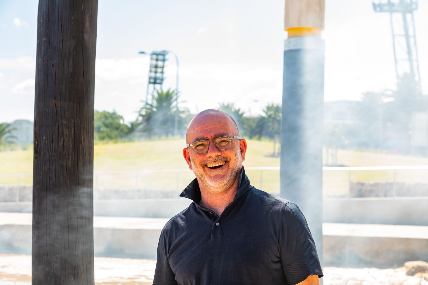 Smiling bald man with glasses and a dark polo shirt standing between two columns in a sunny public park.