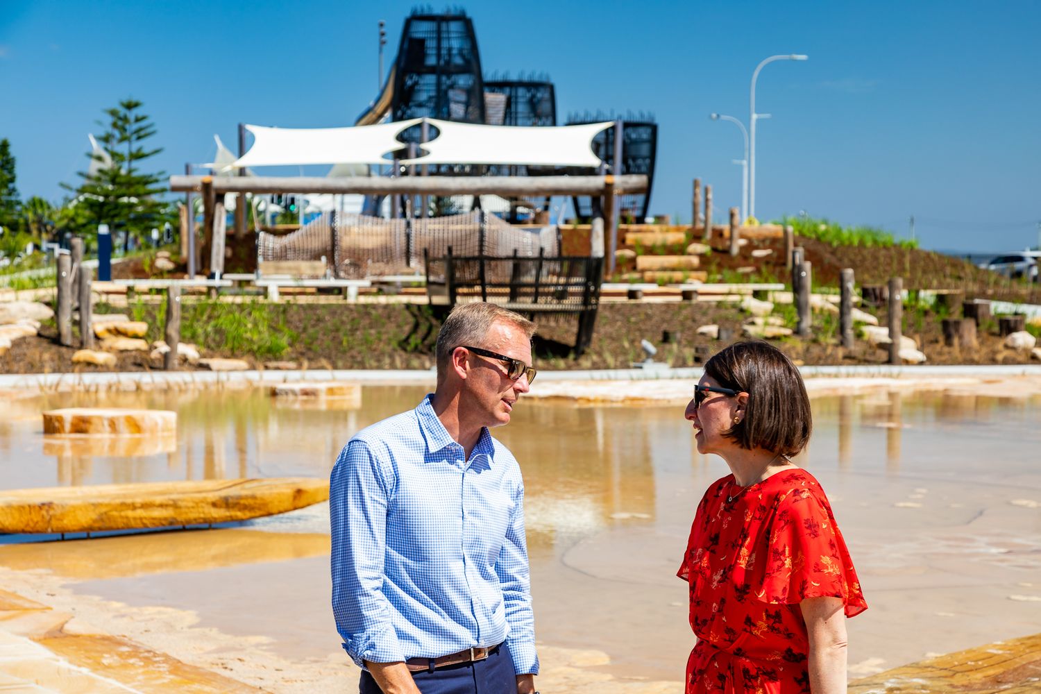 A man in a blue shirt and a woman in a red dress talk beside shallow water, with play structures and shade sails in the background.