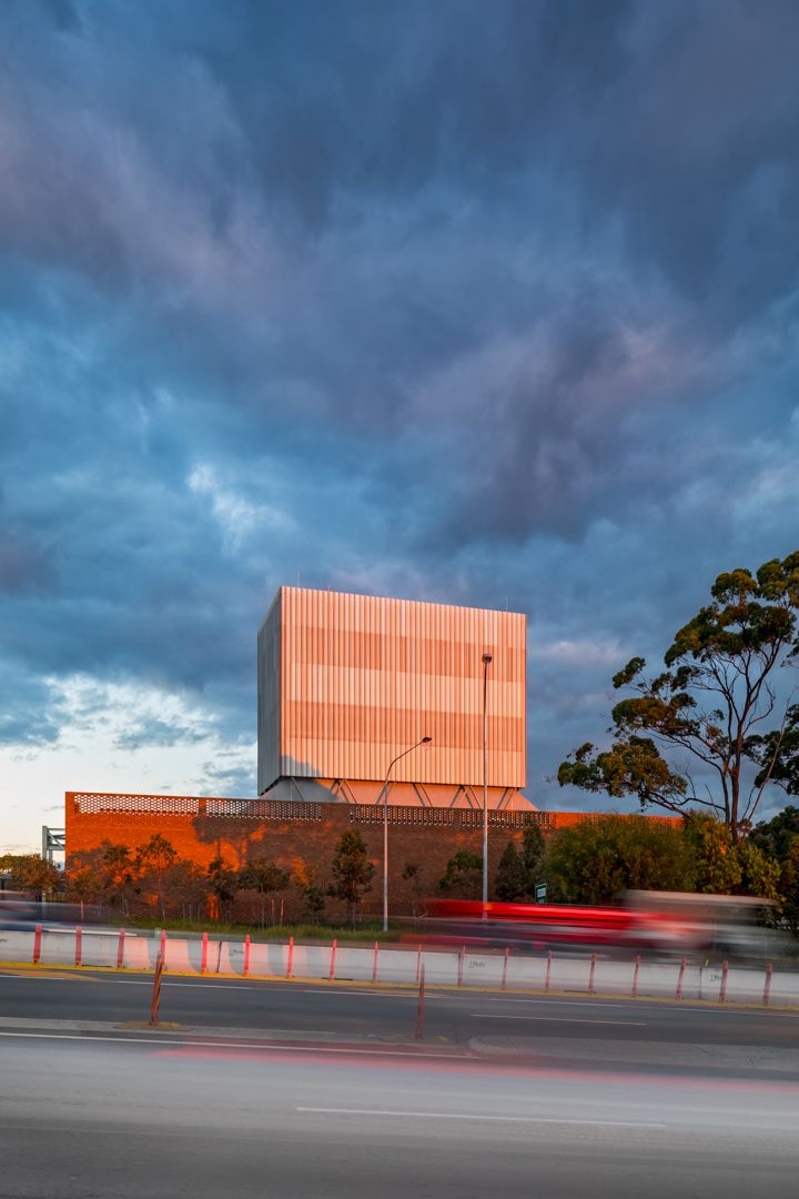 Modern copper-clad building with a vertical louvered facade rises beside a busy road under a dramatic blue stormy sky at dusk.