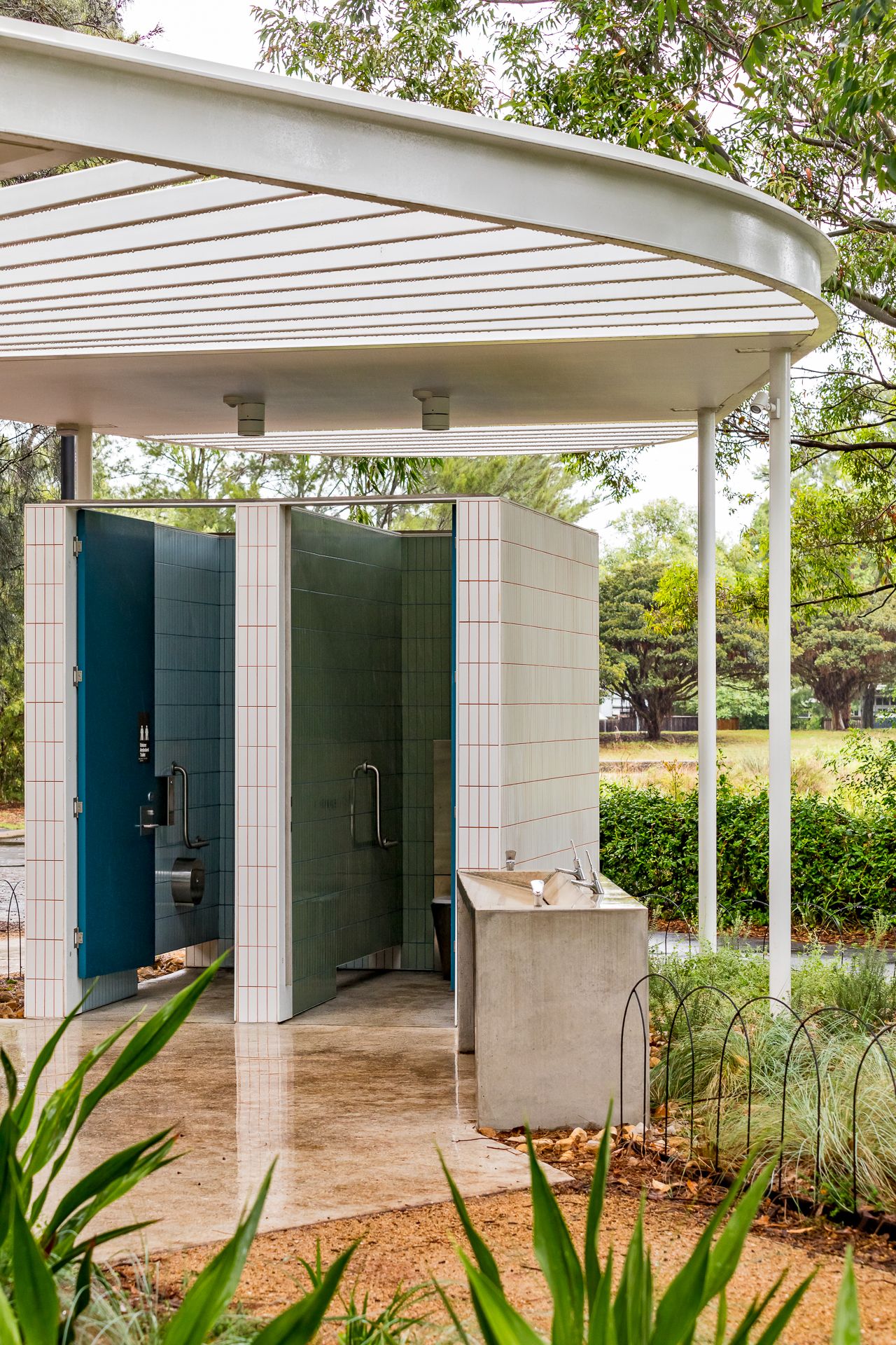 Open-air public restroom with teal doors, white tiles, slim columns and a concrete handwashing trough among greenery.