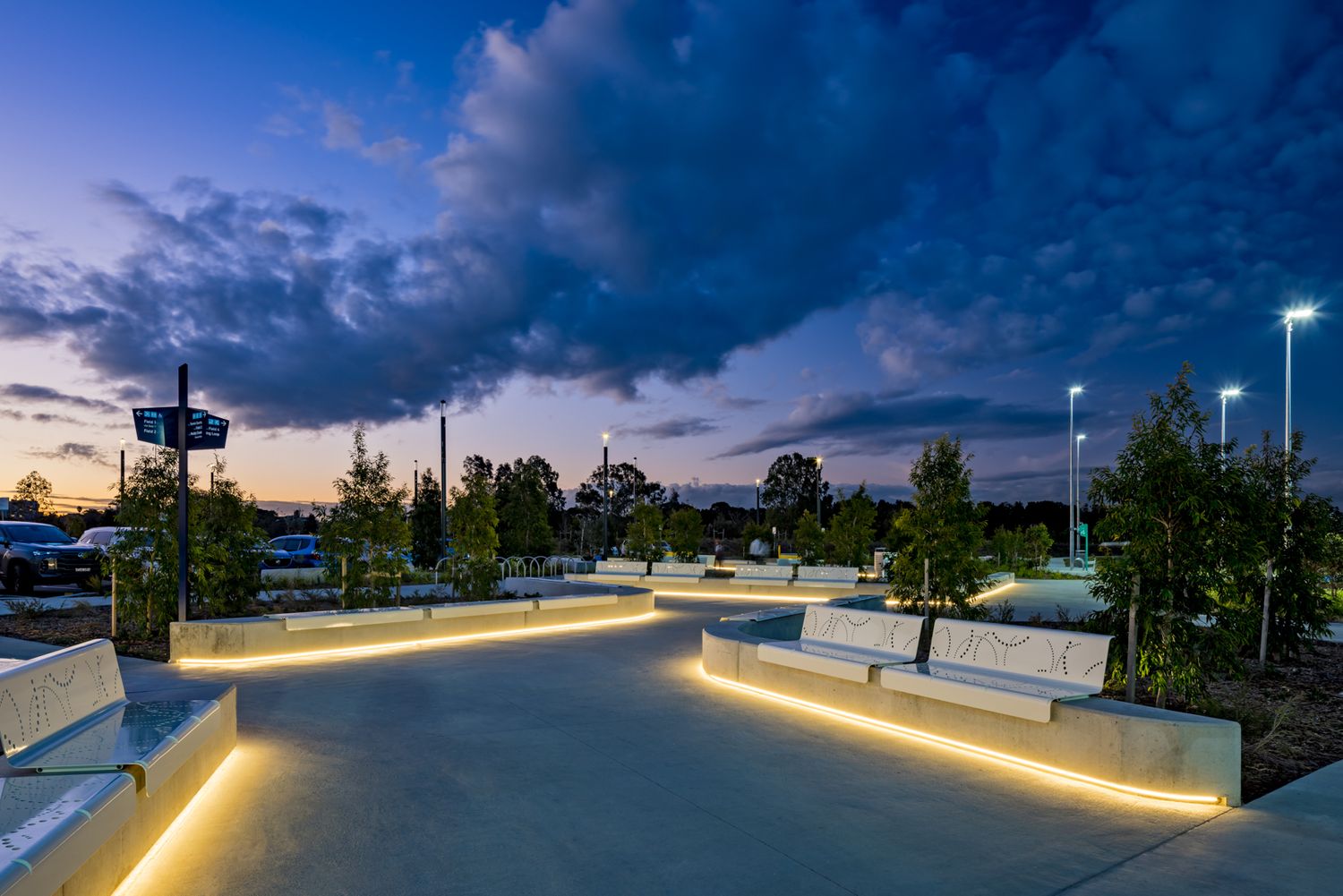 Illuminated seating and curved paths in a modern urban park at dusk under a dramatic blue cloudy sky.