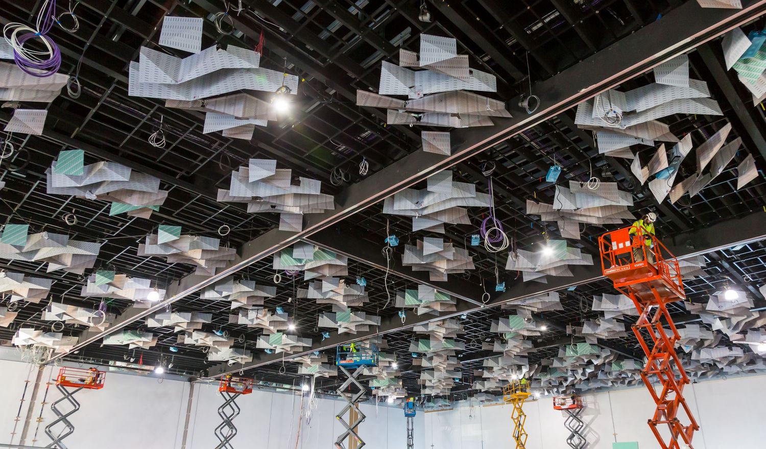 Suspended acoustic ceiling panels in teal and white above scissor lifts inside a building under construction.