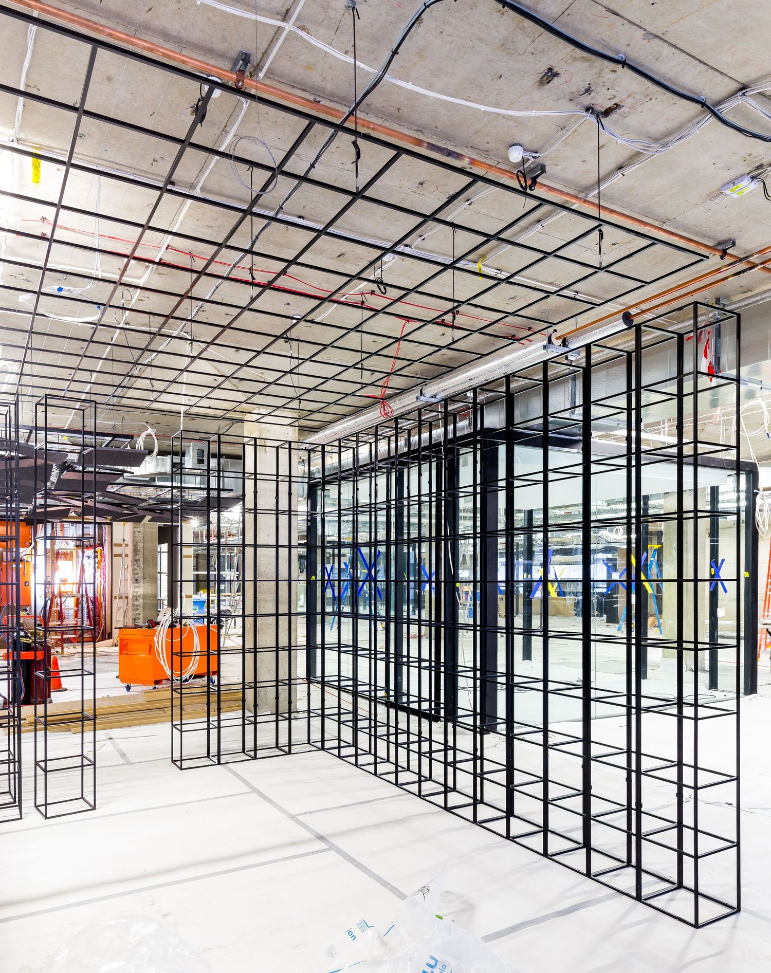 Black steel grid framework structures stand in an unfinished interior space with exposed concrete ceiling and copper pipes.