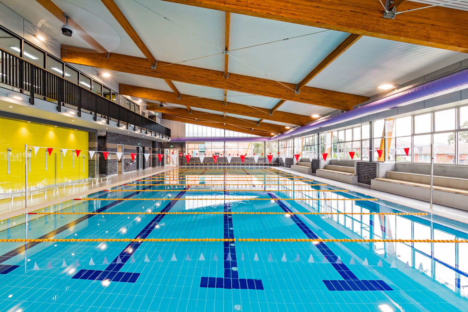 Indoor swimming pool with blue lap lanes, timber ceiling beams, large windows and a yellow feature wall in a leisure centre.