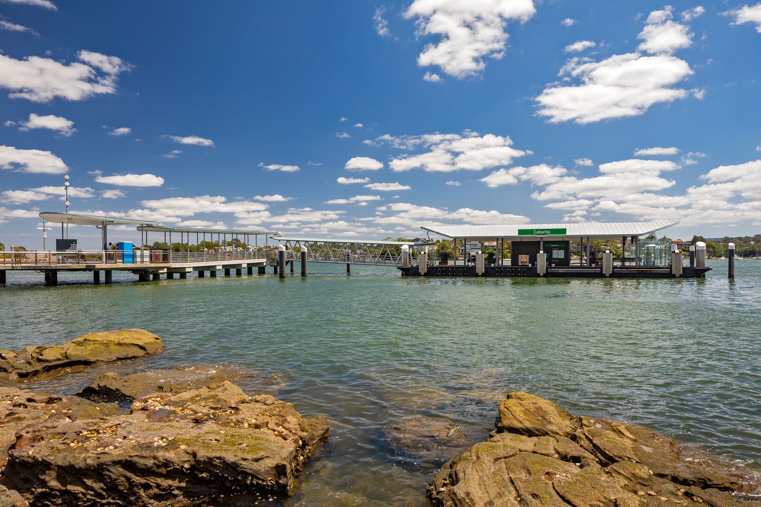 Rocky shoreline in the foreground with a ferry wharf and long jetty across the harbour under a blue sky with white clouds.