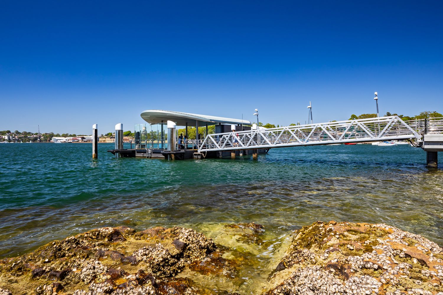 Ferry wharf with a curved roof pontoon and white gangway beside rocky shoreline, under a vivid blue sky over Sydney Harbour.