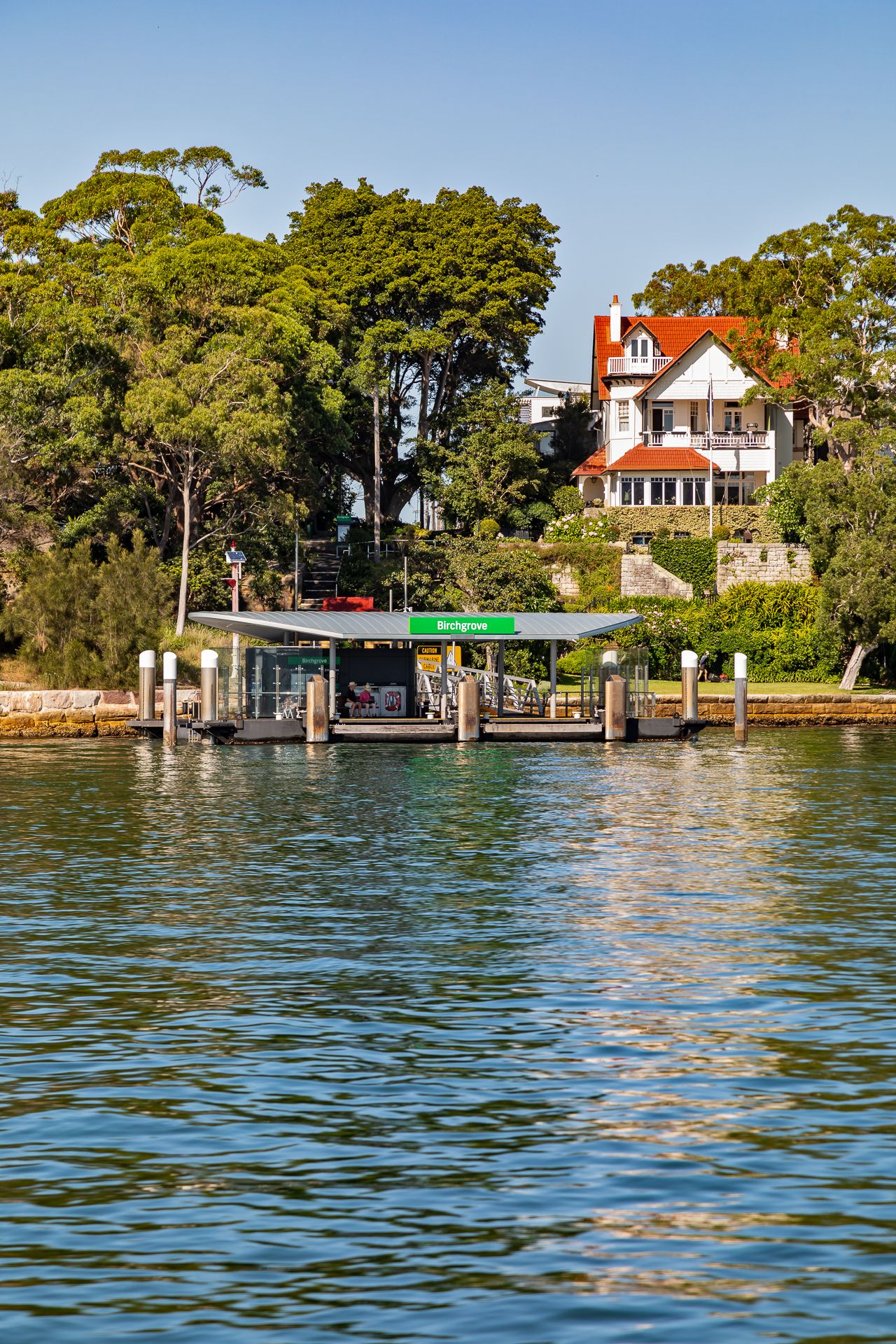 Ferry wharf with a green name sign sits on the harbour water in front of tall trees and a red-roofed heritage house on a sunny day.