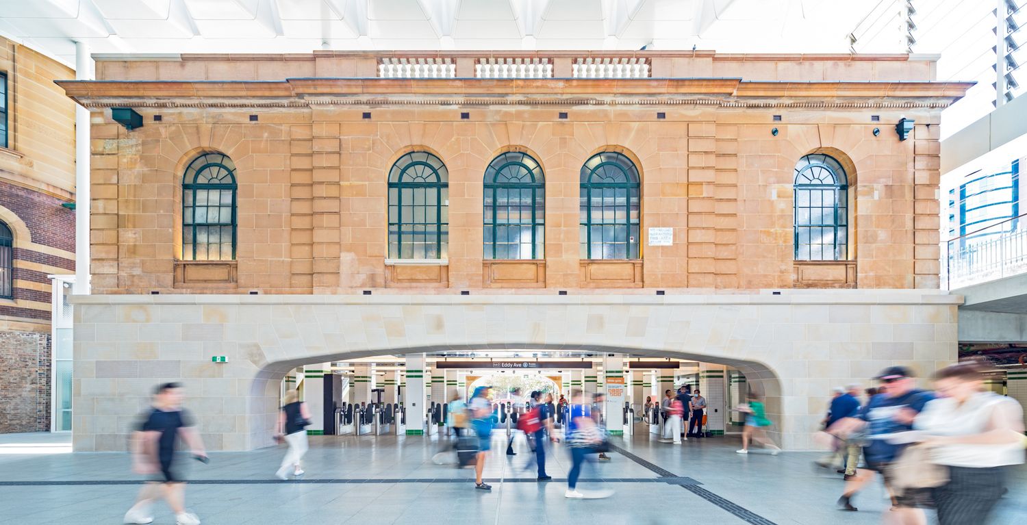 Historic brick building with arched windows rising above a bright modern concourse where blurred pedestrians walk through.