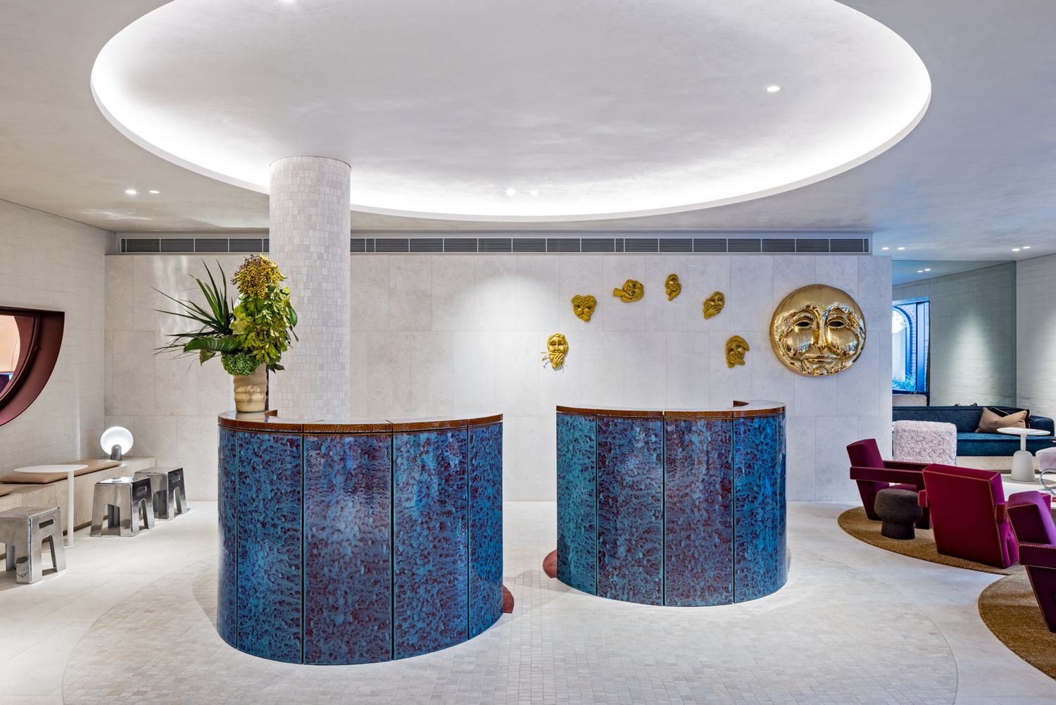 Hotel reception with two curved blue tiled desks beneath a circular ceiling light, gold wall masks, a potted plant and red armchairs.