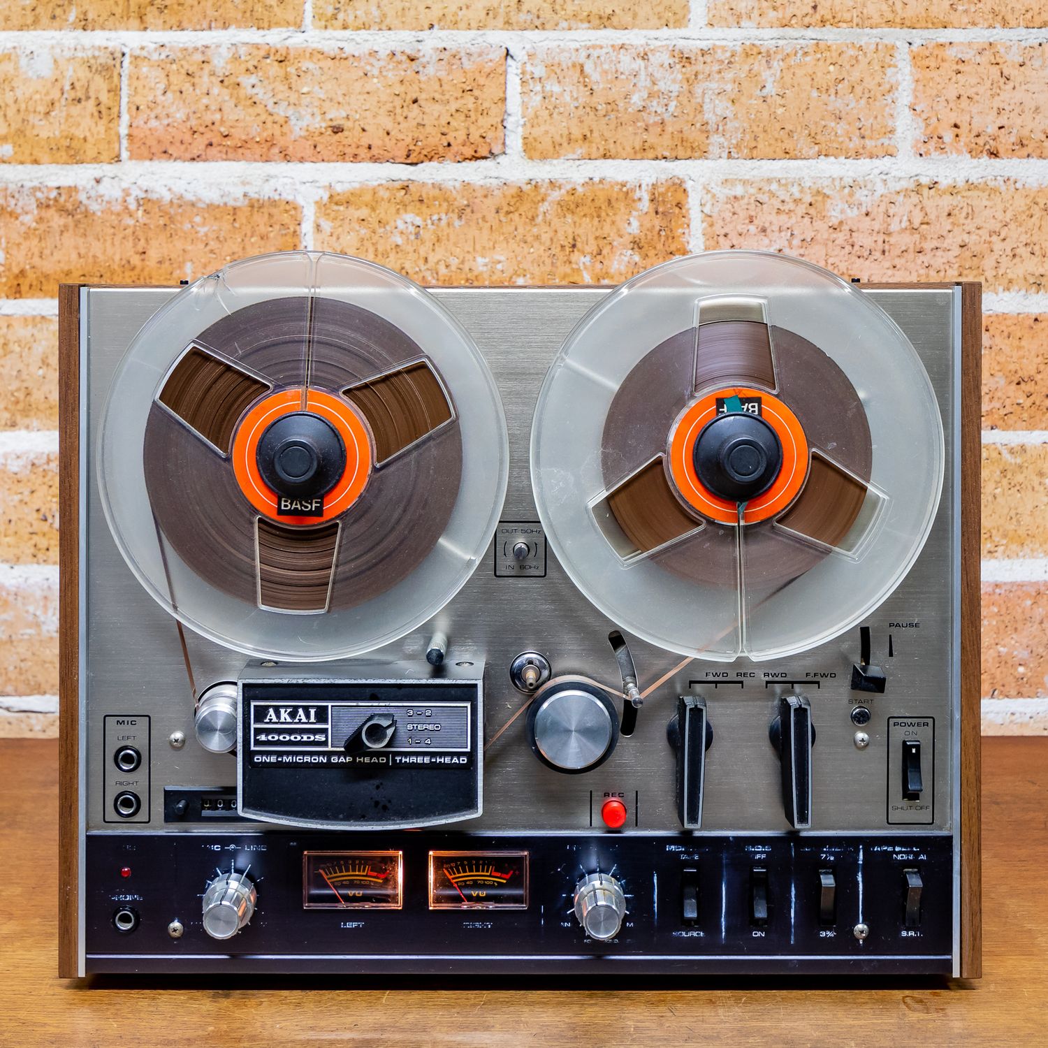 Vintage Akai reel-to-reel tape recorder with orange reels and analog controls in front of a rustic brick wall.