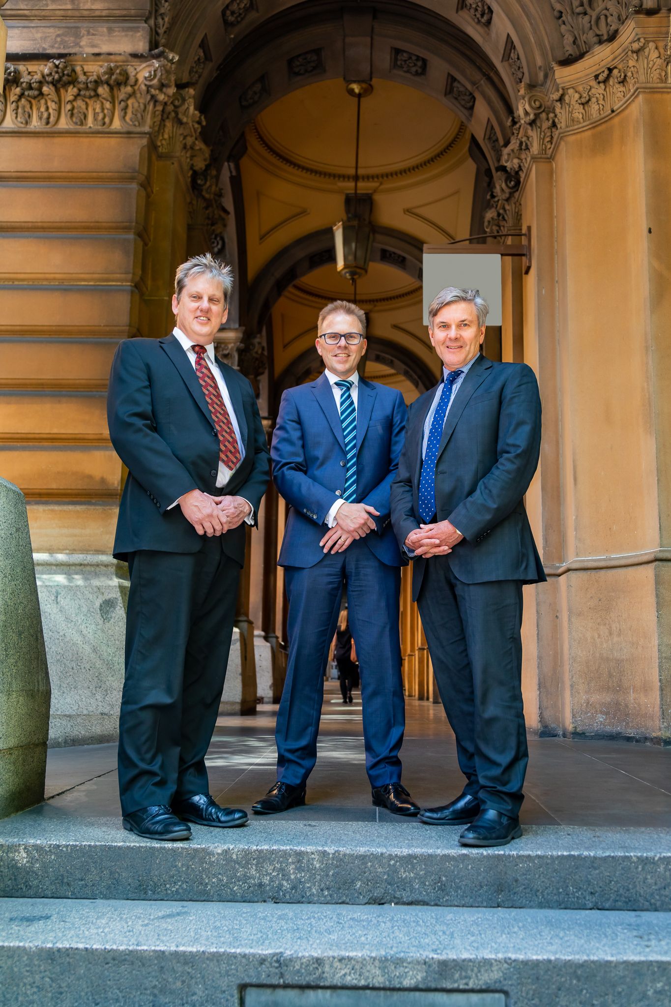 Three men in dark business suits stand confidently on stone steps in front of a grand sandstone building archway.