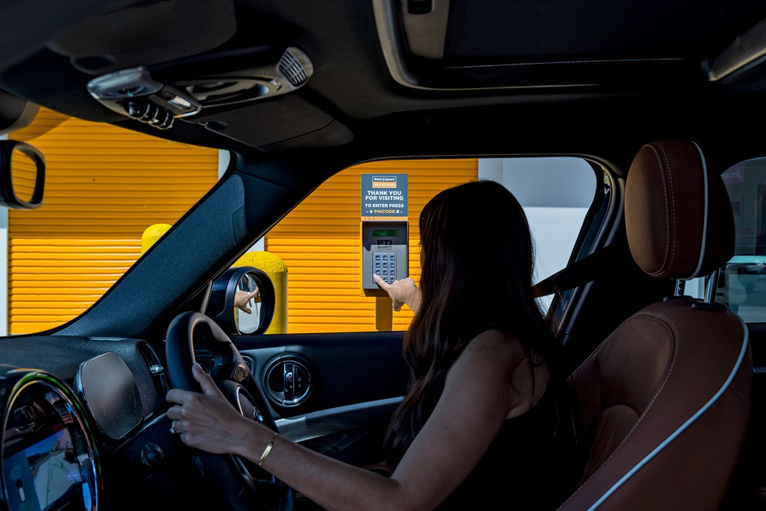 Woman with long dark hair sits in the driver's seat of a car, reaching toward a drive-through window with a yellow wall.