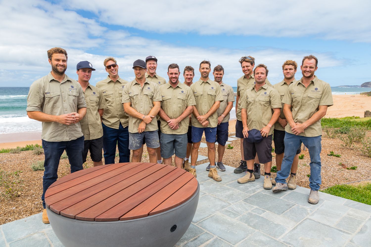 Group of smiling men in matching khaki work shirts and shorts posing outdoors beside a round timber-topped fire pit near a sandy beach.