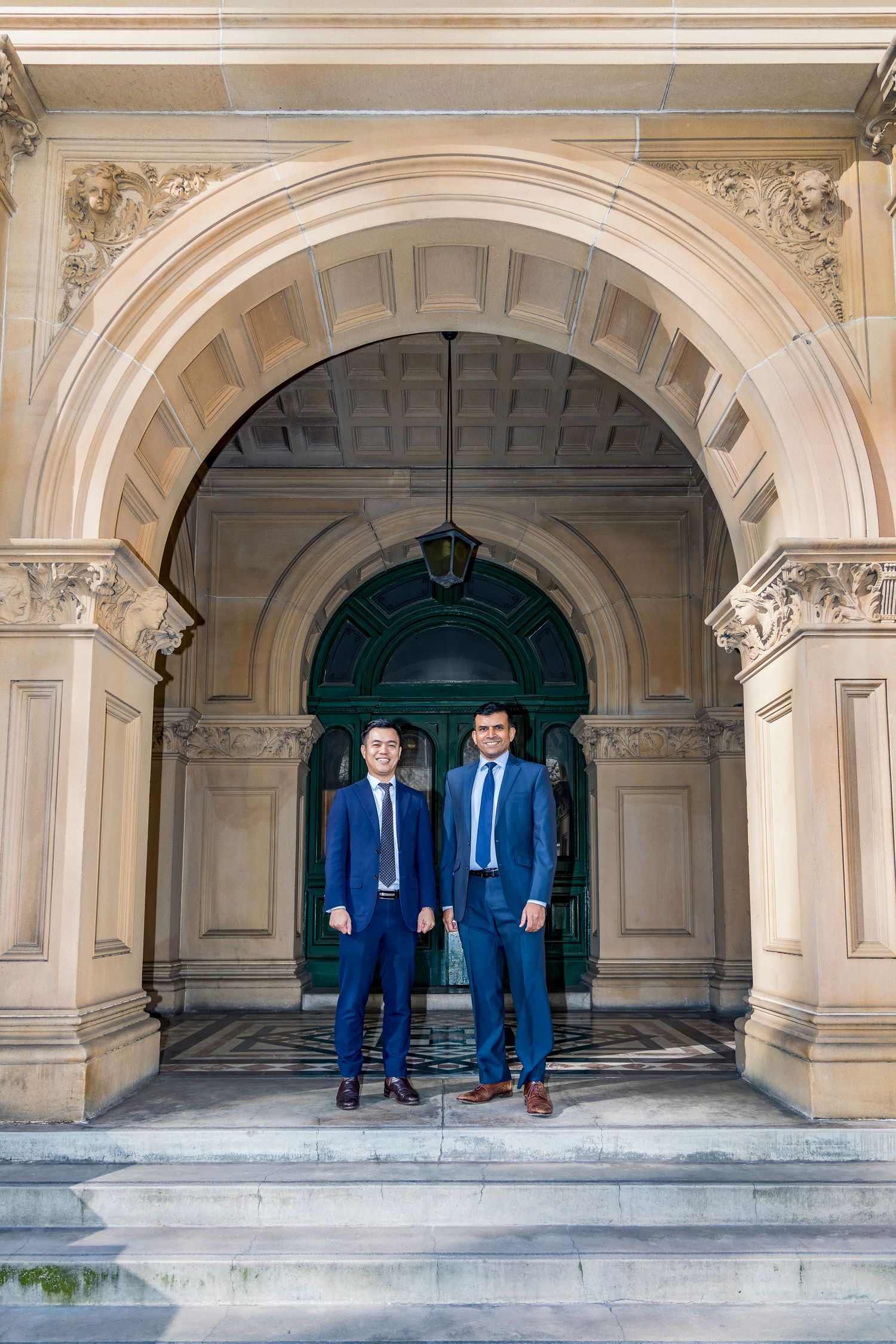 Two men in navy suits and ties stand side by side beneath an ornate sandstone archway at the entrance of a historic building.