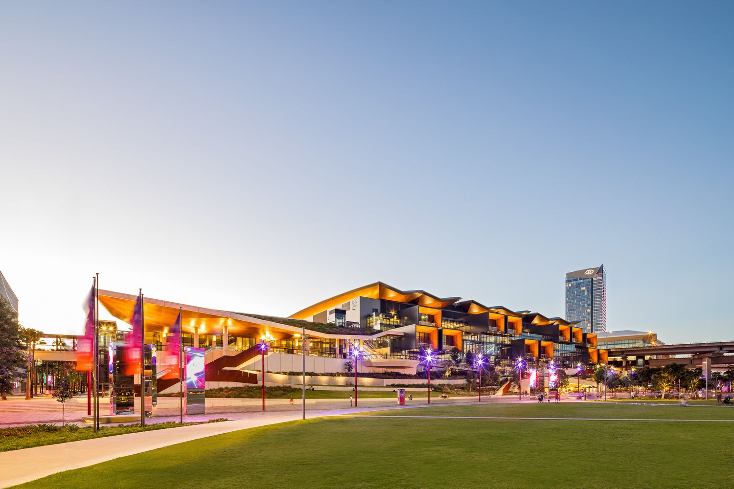 Colorful event centre with curved timber-clad roofs and illuminated flags beside a green lawn at dusk, with a high-rise tower behind.