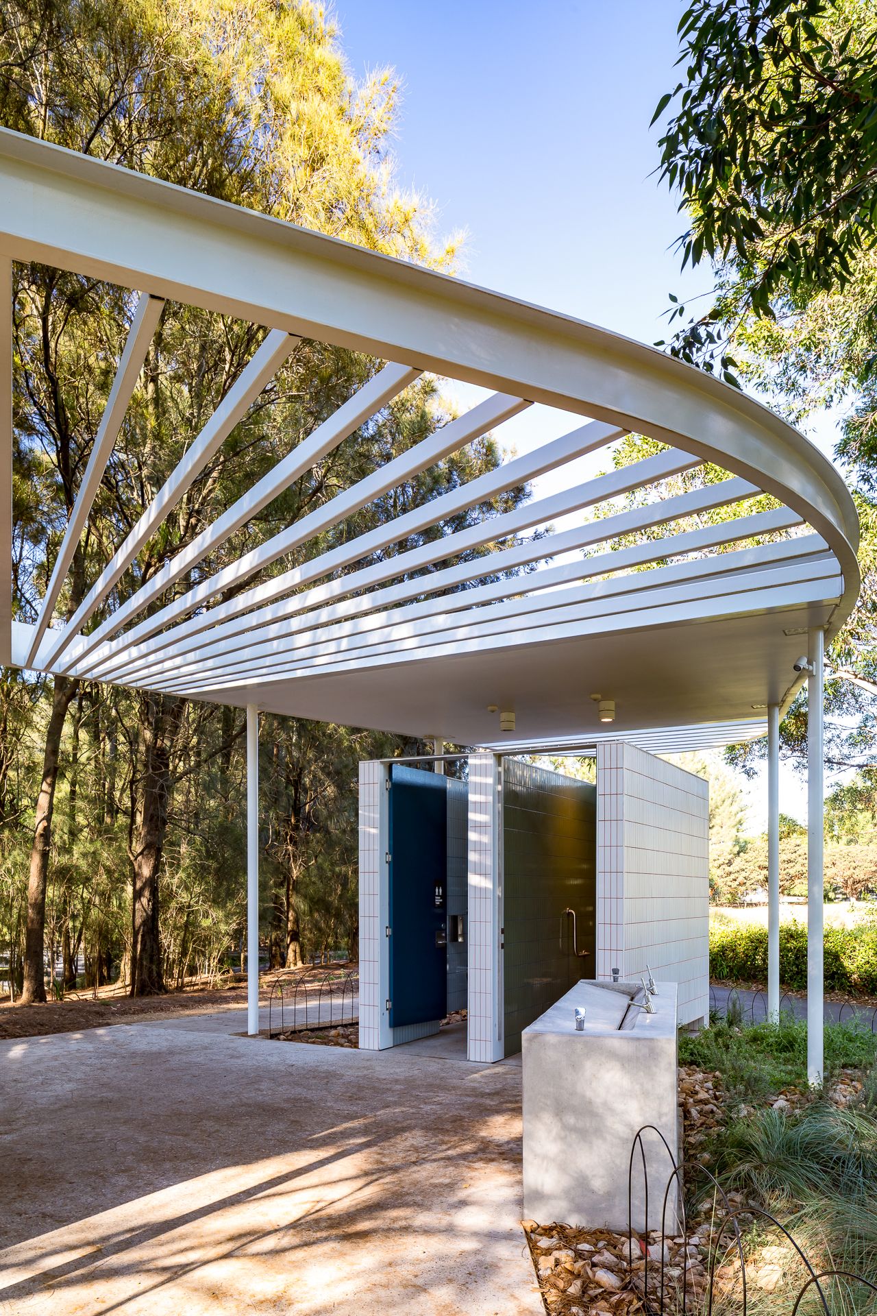 Curved white slatted roof canopy over blue restroom doors and a drinking fountain in a modern park building surrounded by tall trees.
