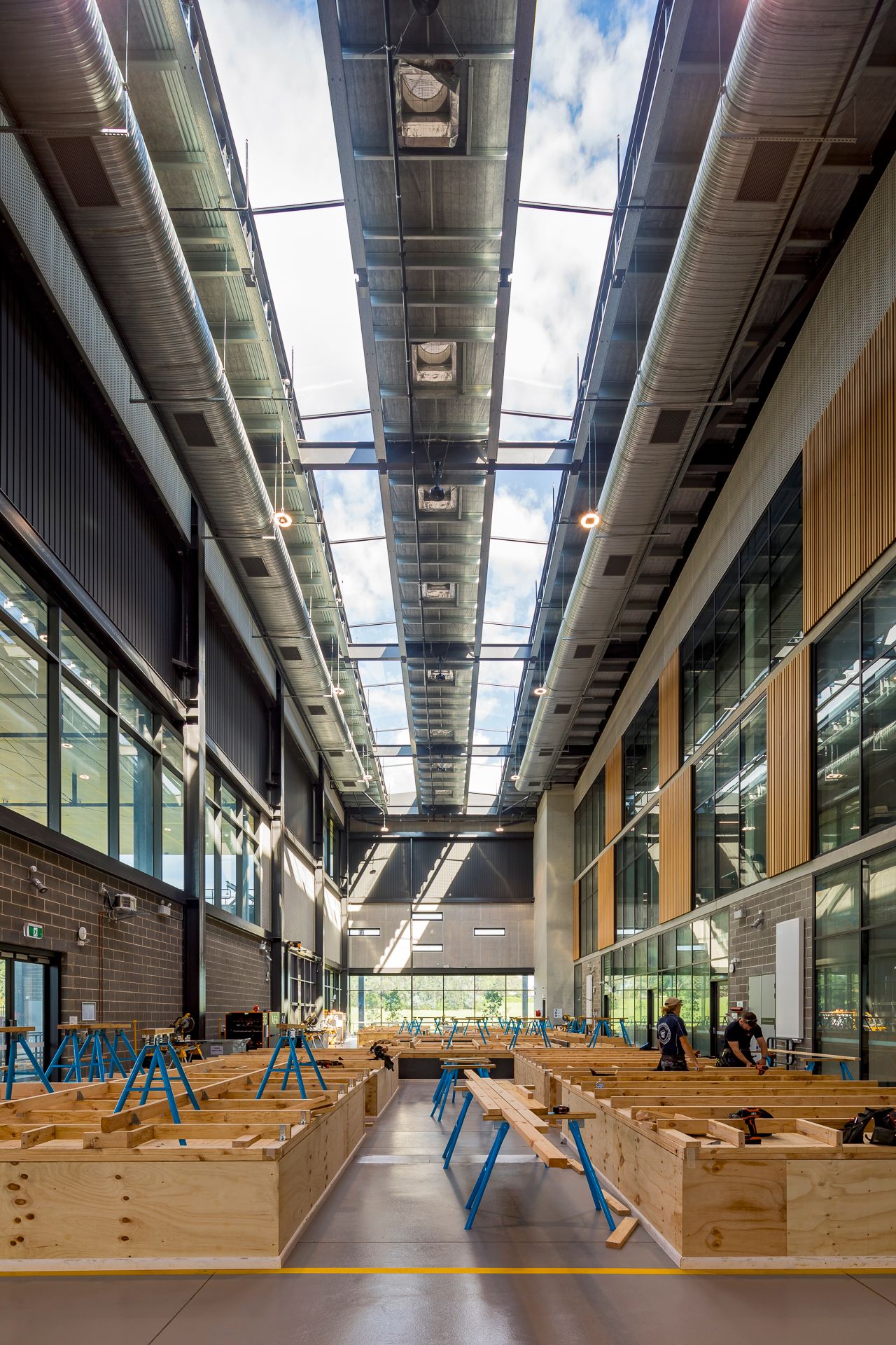 Tall skylit TAFE carpentry workshop with timber workbenches, blue sawhorses, exposed ducting and students working below timber-clad walls.