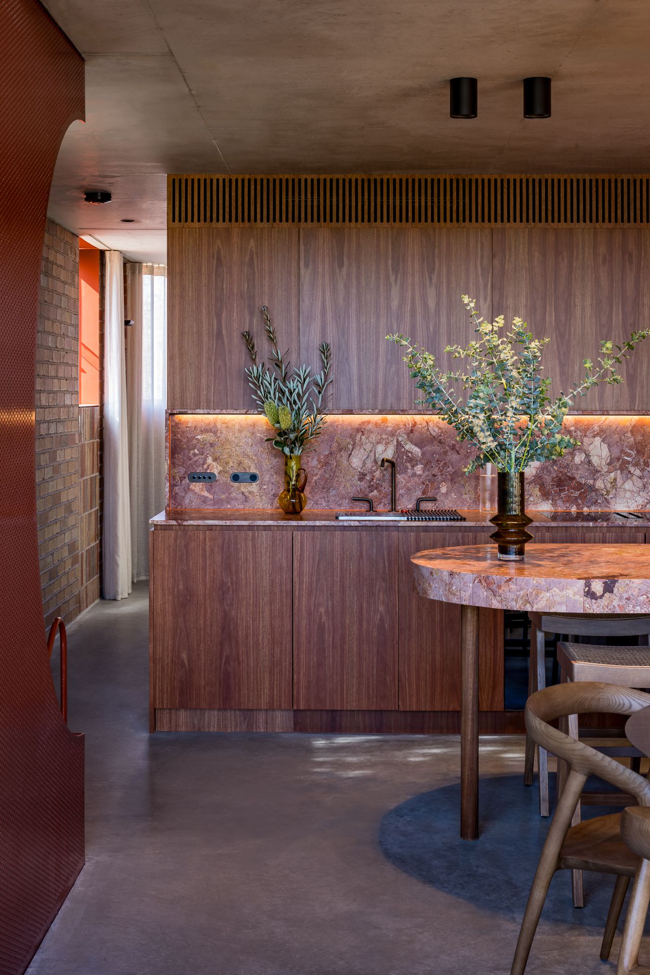 Modern kitchen with walnut cabinetry, pink marble backsplash, warm under-cabinet lighting, a round marble table and eucalyptus in vases.