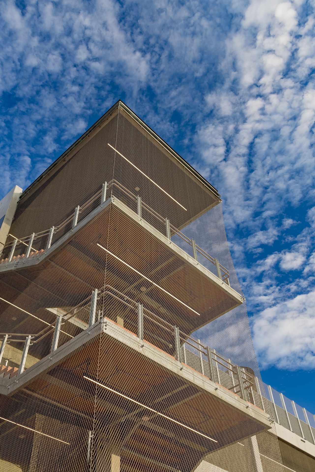 Angular multi-story building with open balconies and railings wrapped in steel mesh, set against a bright blue sky with wispy clouds.