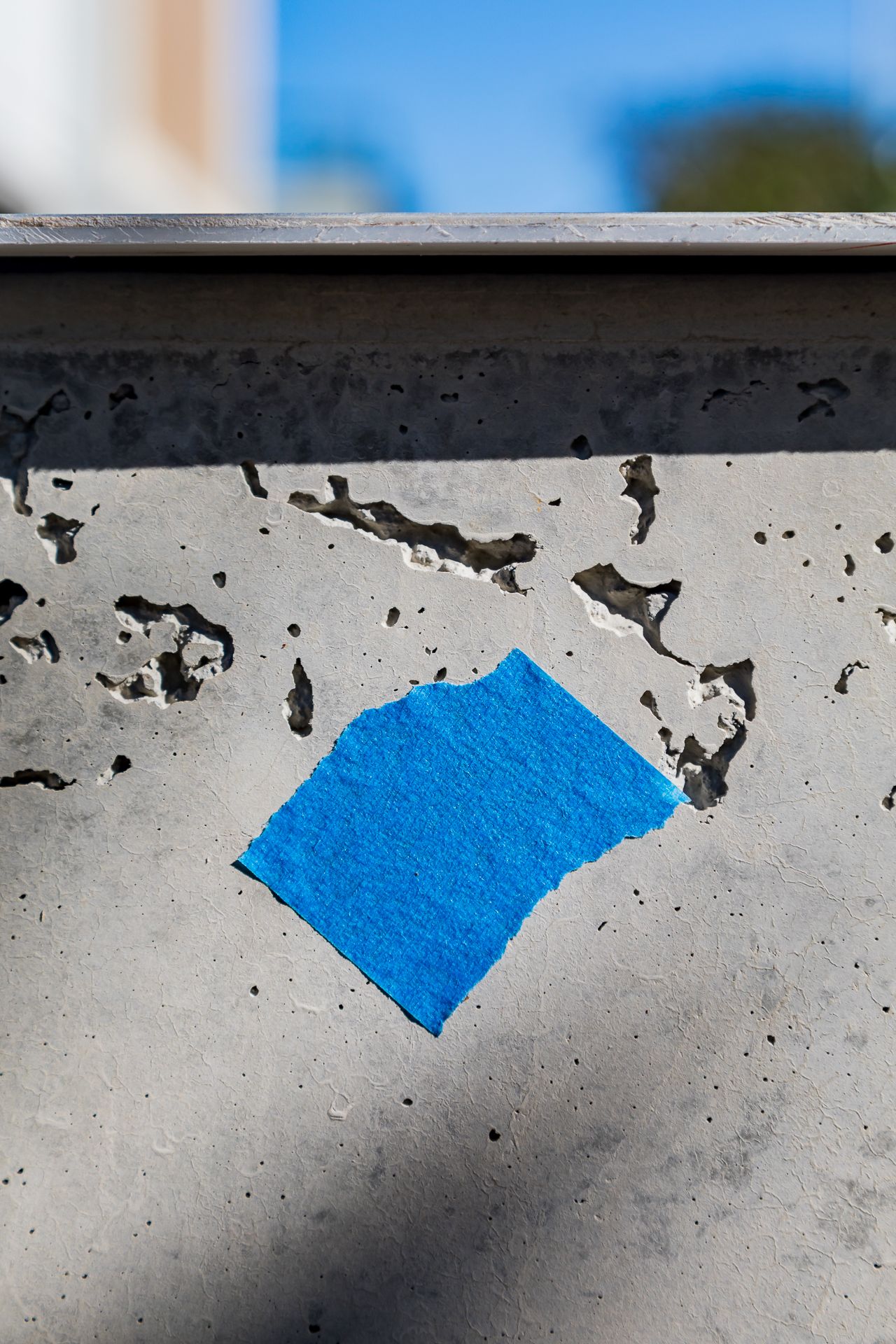 Torn piece of blue masking tape stuck to a weathered concrete wall with pitted holes and chipped, crumbling surface.
