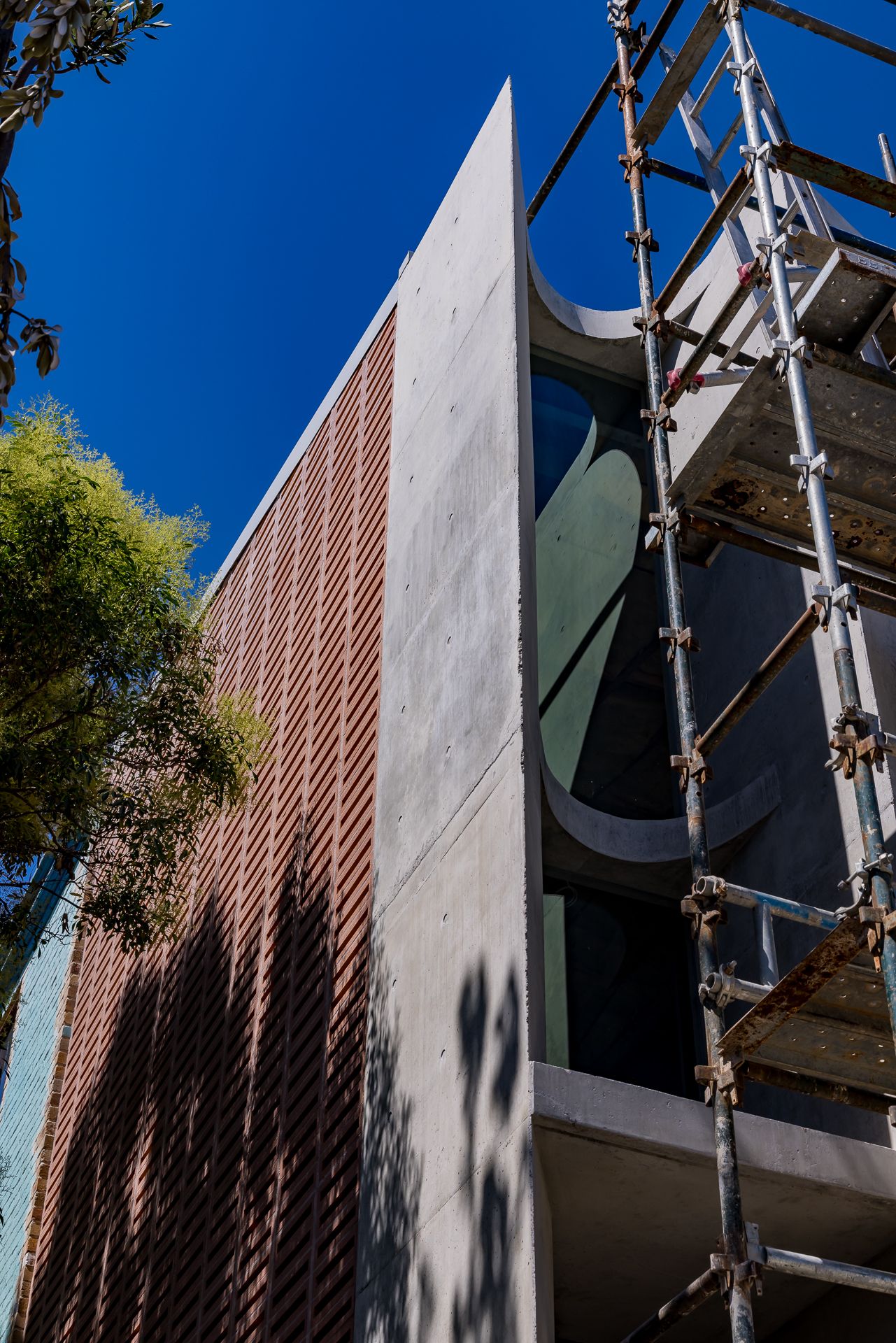 Modern building exterior with a board-formed concrete wall, terracotta brick screen and curved glazing, with scaffolding against a blue sky.