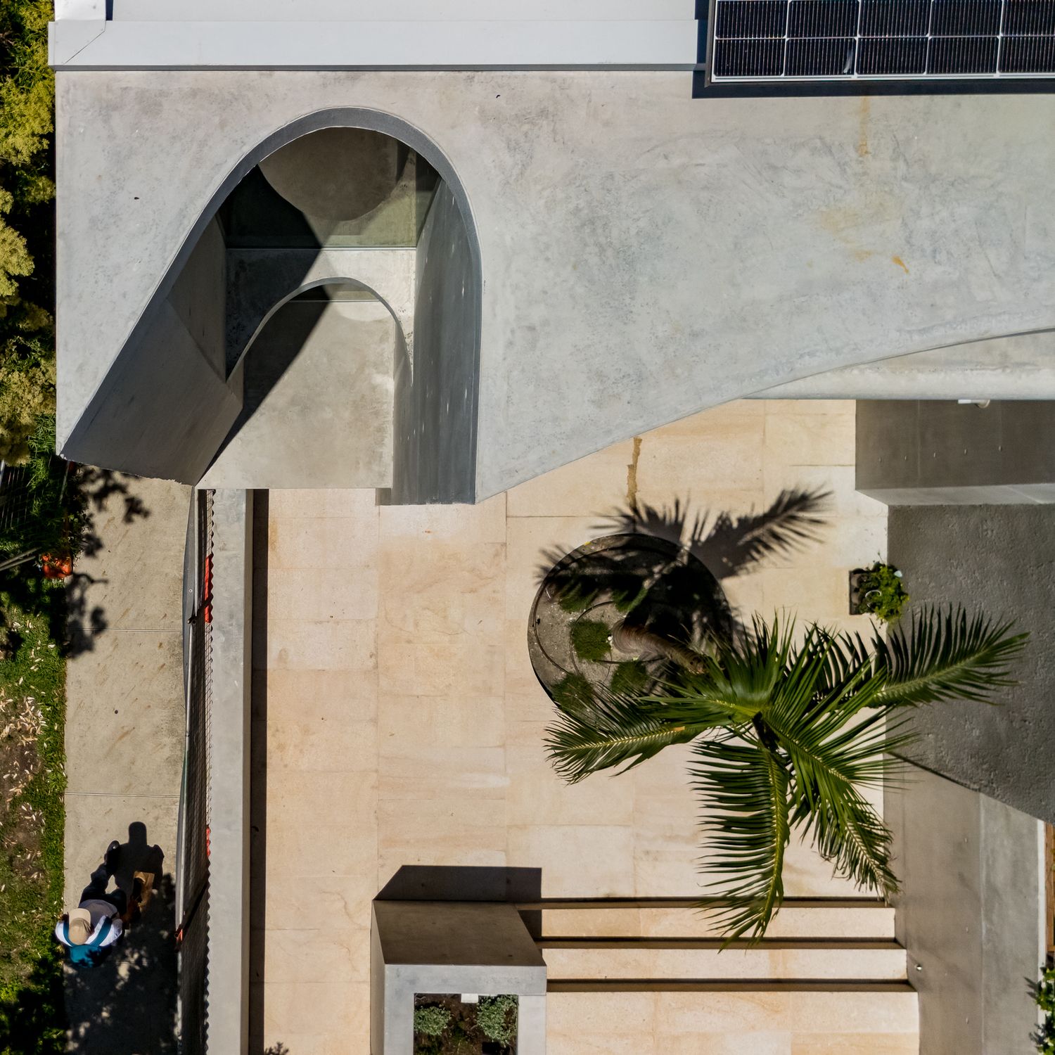 Aerial view of a concrete building with arched opening, solar panel, palm tree and sandstone paved courtyard.