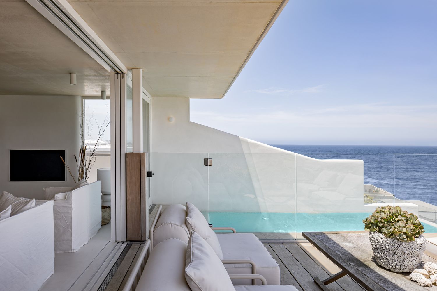 Terrace with outdoor lounge seating beside a glass-fronted plunge pool overlooking the ocean from a modern white home.