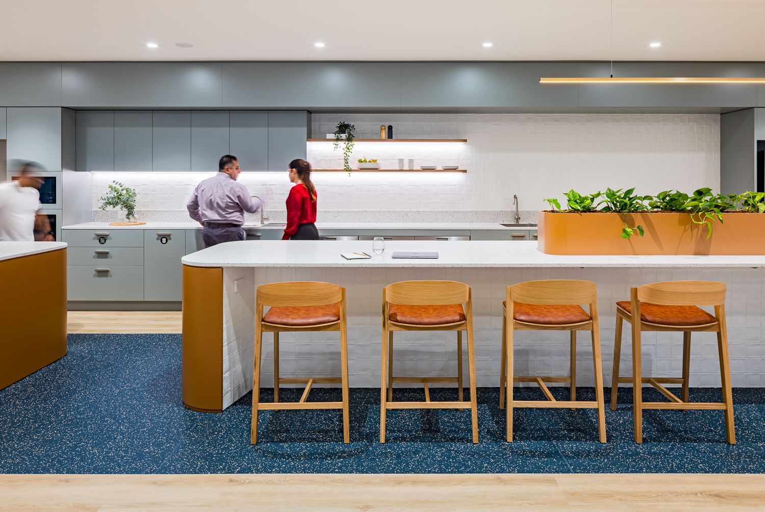 Modern office kitchen with a long white island, orange timber barstools, blue speckled flooring and two colleagues chatting at the counter.