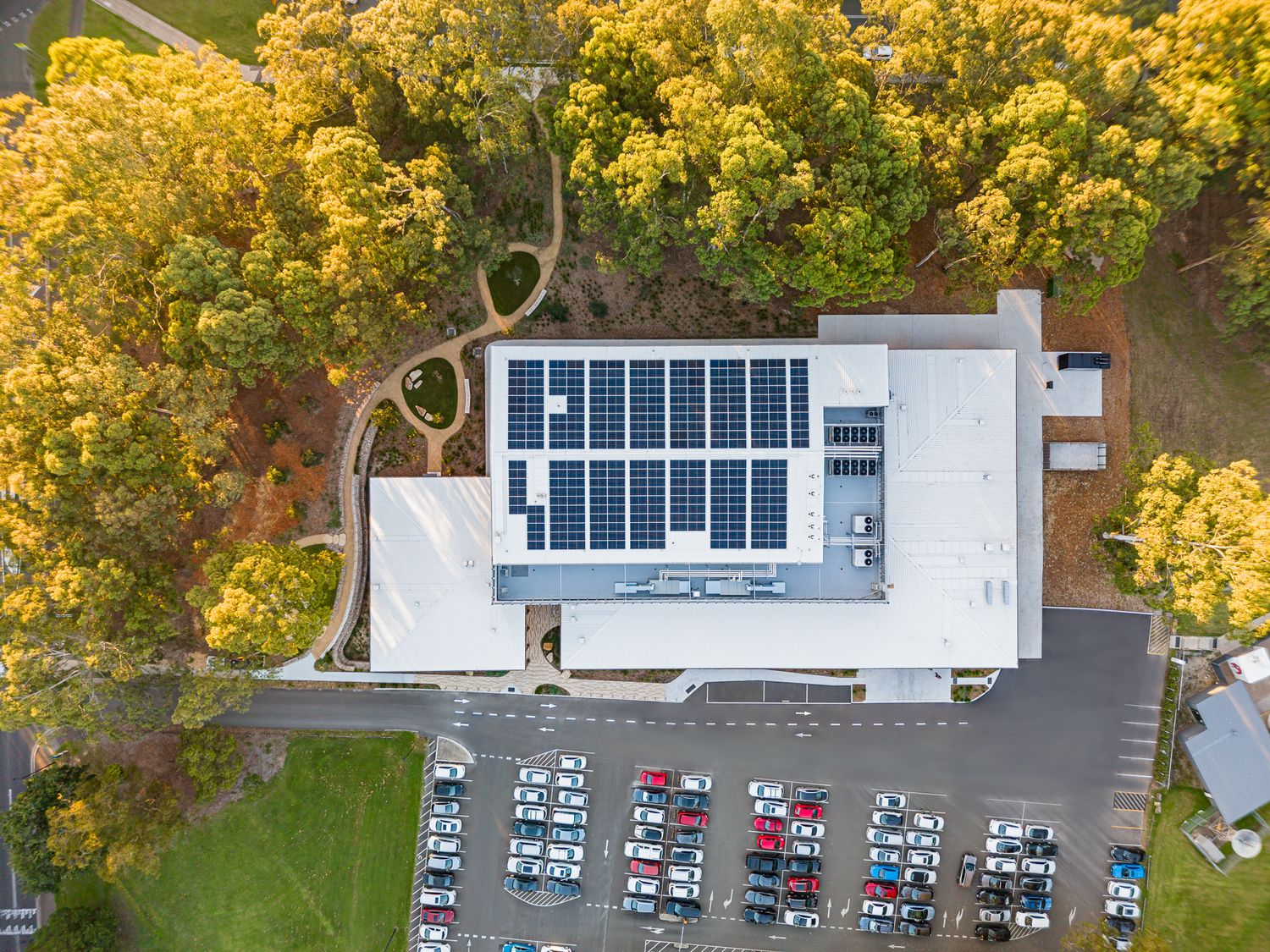 Top-down drone view of a facility roof covered in solar panels beside a car park and autumn-colored trees.