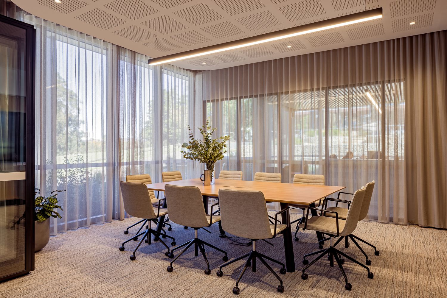 Modern meeting room with a long timber table, cream swivel chairs, sheer curtains and large windows overlooking trees.